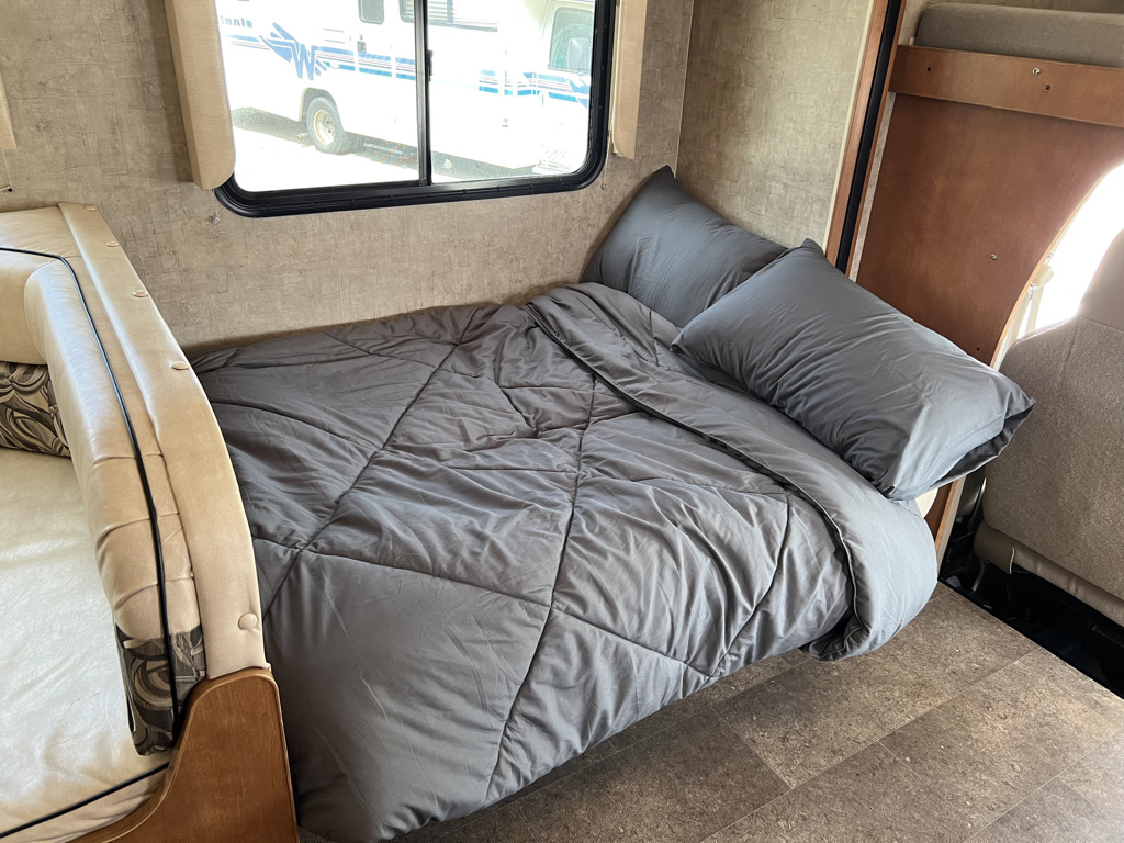 An RV rental interior features a cozy, grey-comforter bed with pillows, next to a window. Ideal sleeping space for travelers.. Winnebago Minnie Winnie 2015