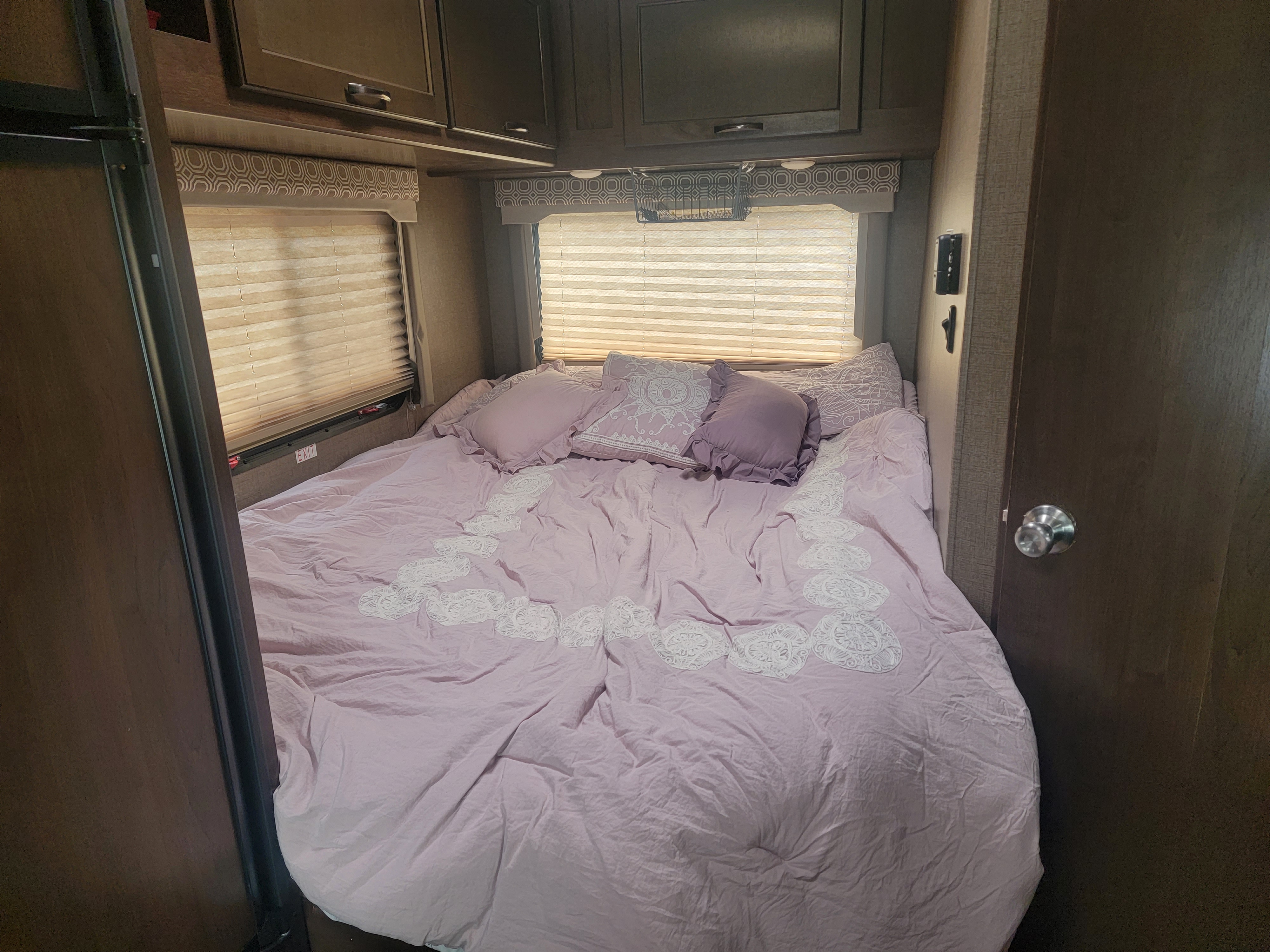 Cozy RV rental bedroom featuring a lavender bed with decorative pillows, window blinds, overhead cabinets, and an emergency exit.. Thor Four Winds 2020
