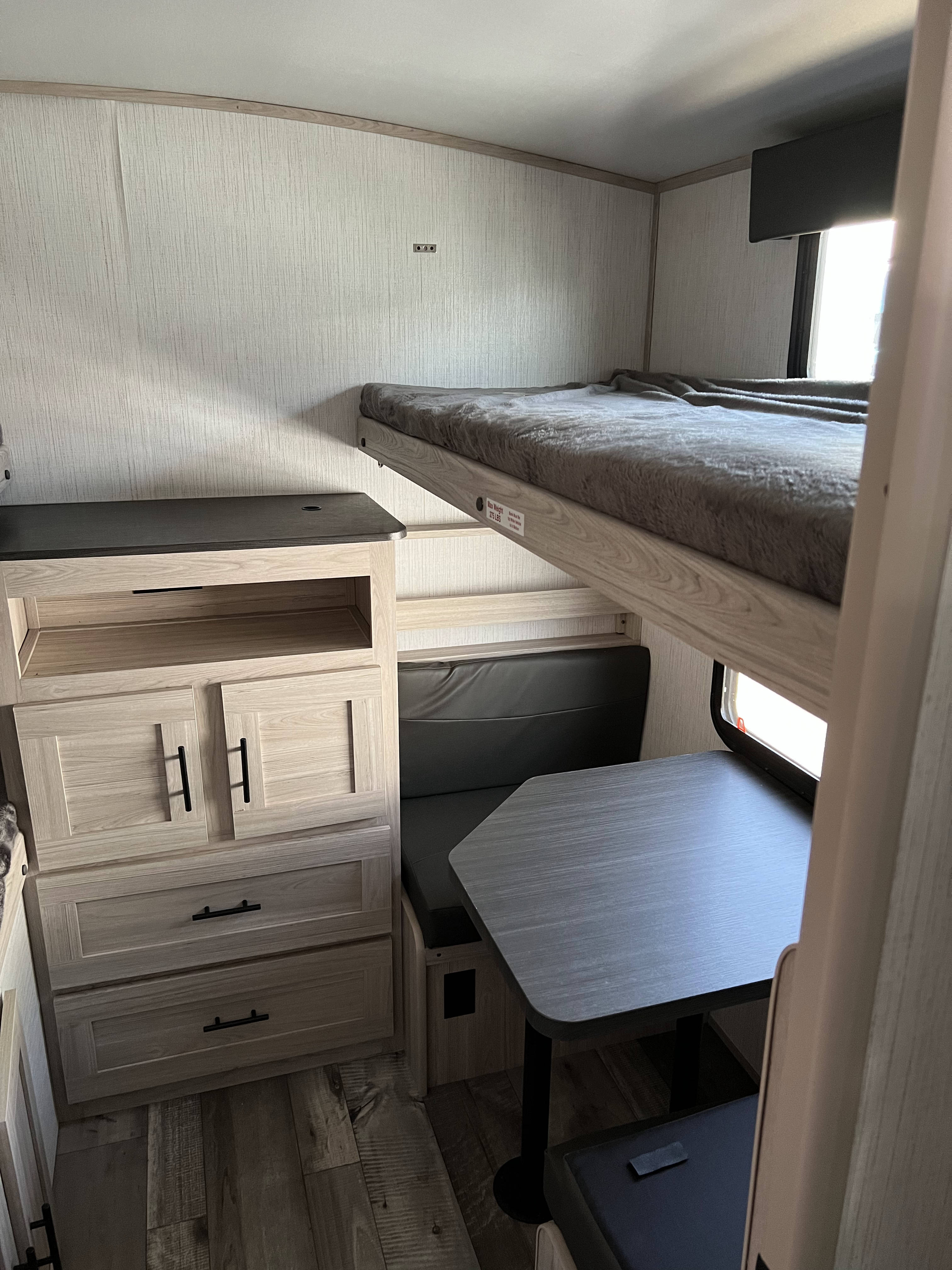 An RV rental interior with light wood bunk beds (top bunk has gray blanket) above a dark gray dinette with a table and built-in storage.. Heartland RVs Sundance Ultra Lite 2022