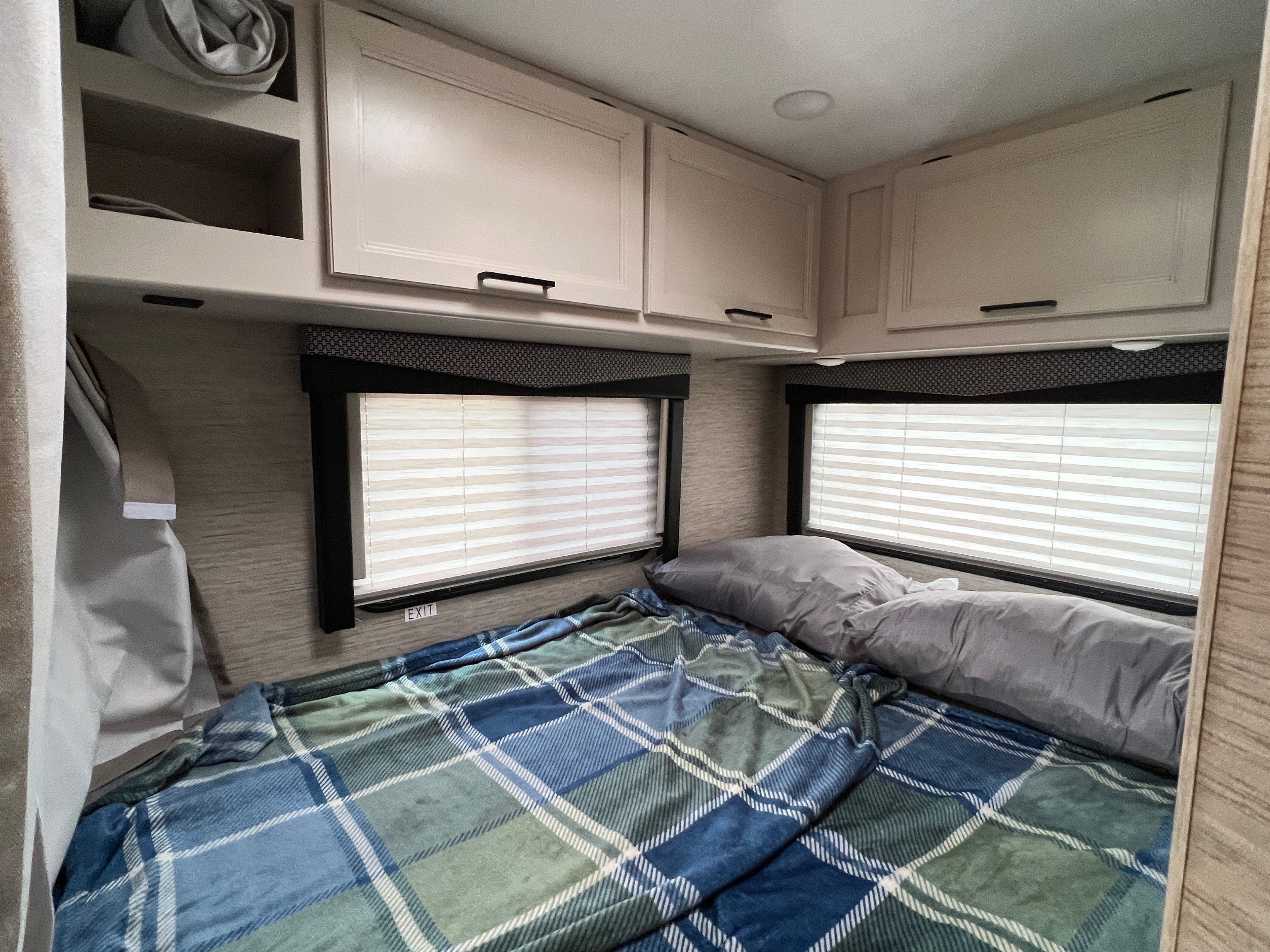 Cozy RV rental bedroom featuring a plaid blanket, gray pillows, windows with blinds, and overhead storage cabinets.. Thor Freedom Elite 2023