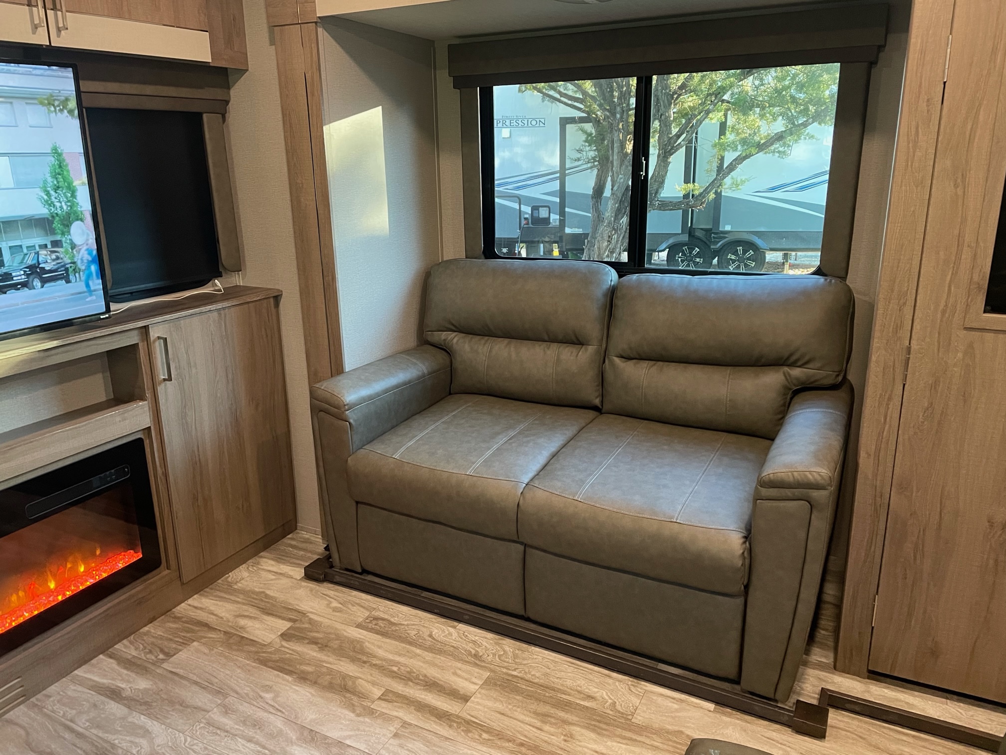 Inside an RV rental: a cozy living area with a gray sofa, TV, and an electric fireplace. Outside, another RV is parked.. Grand Design Imagine RL 3100 2022