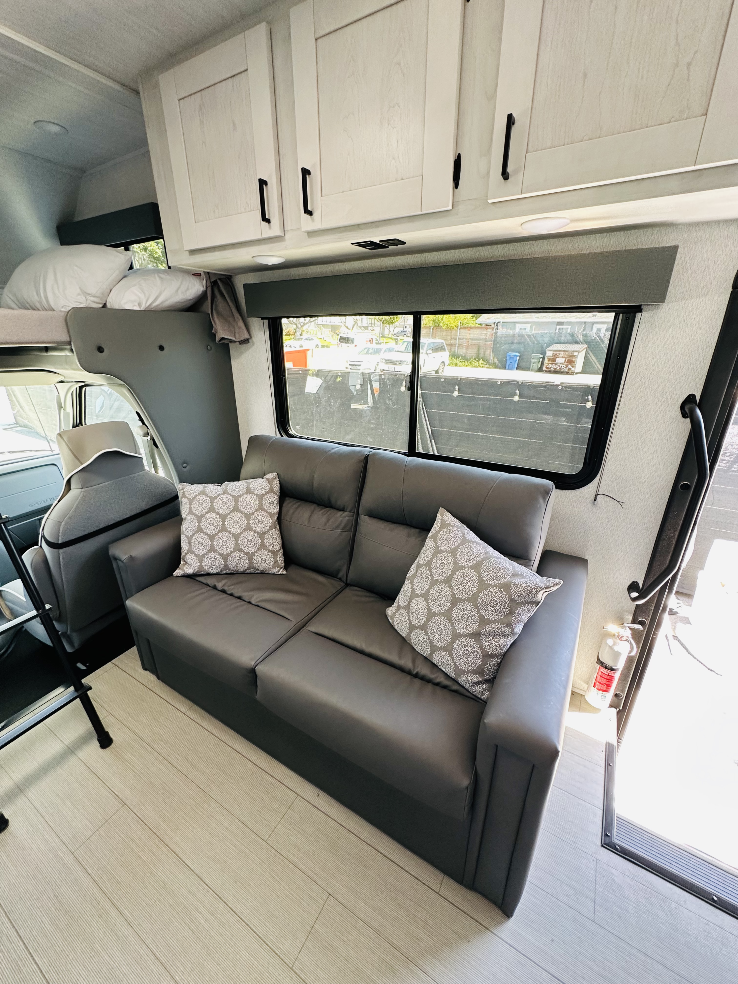 Spacious RV rental interior features a dark gray sofa, light wood-look cabinets, bunk bed, window, and plank flooring.. East to West Entrada 2023