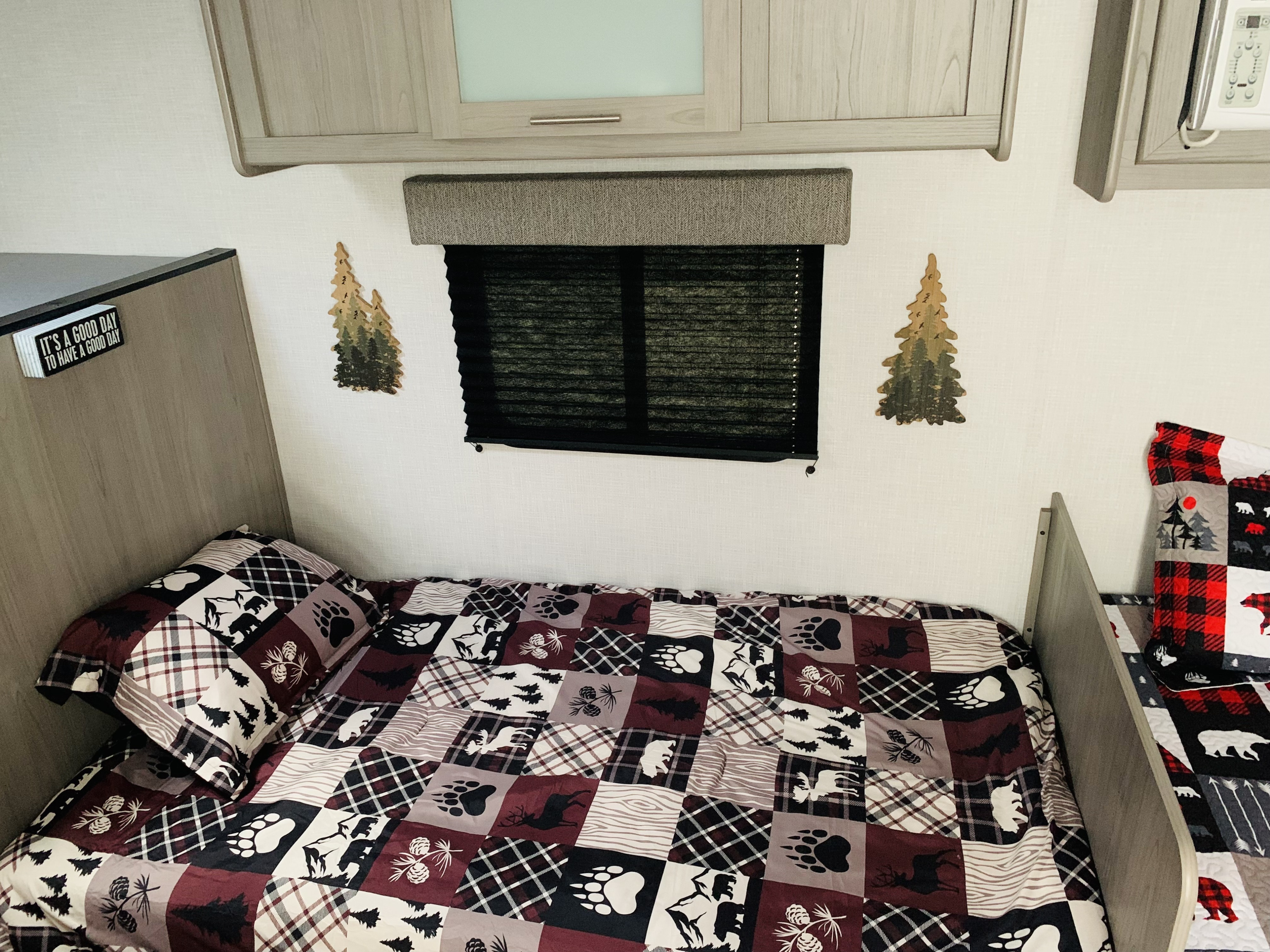 Inside an RV rental, a bed with a nature-themed quilt (bears, plaid) and window blinds offers a cozy sleeping area.. Dutchmen Coleman Lantern LT 2023