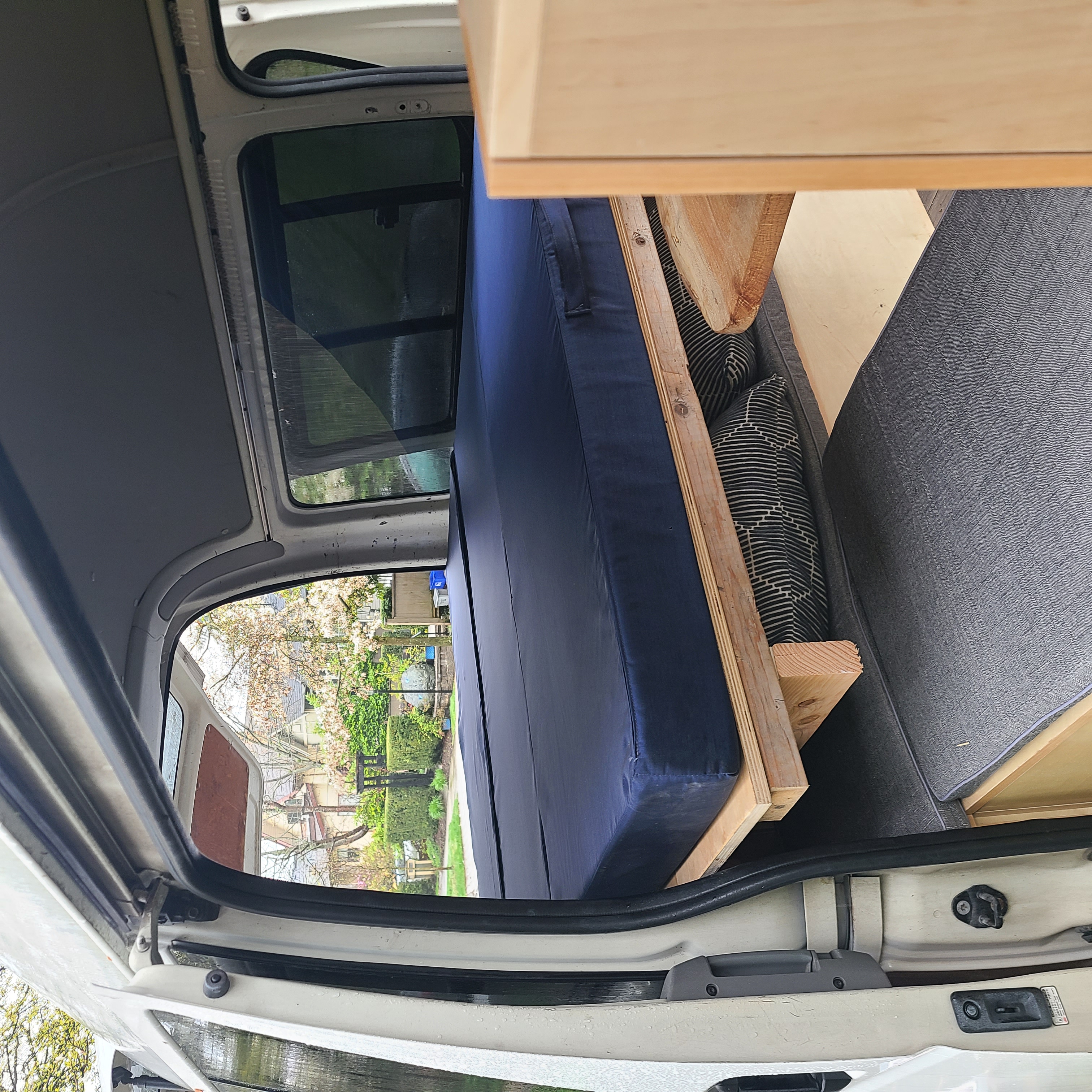 Sideways view inside an rv rental camper van: A blue bed, wooden frame, and grey cushions. Open doors show an outdoor scene.. Toyota Hiace 2006