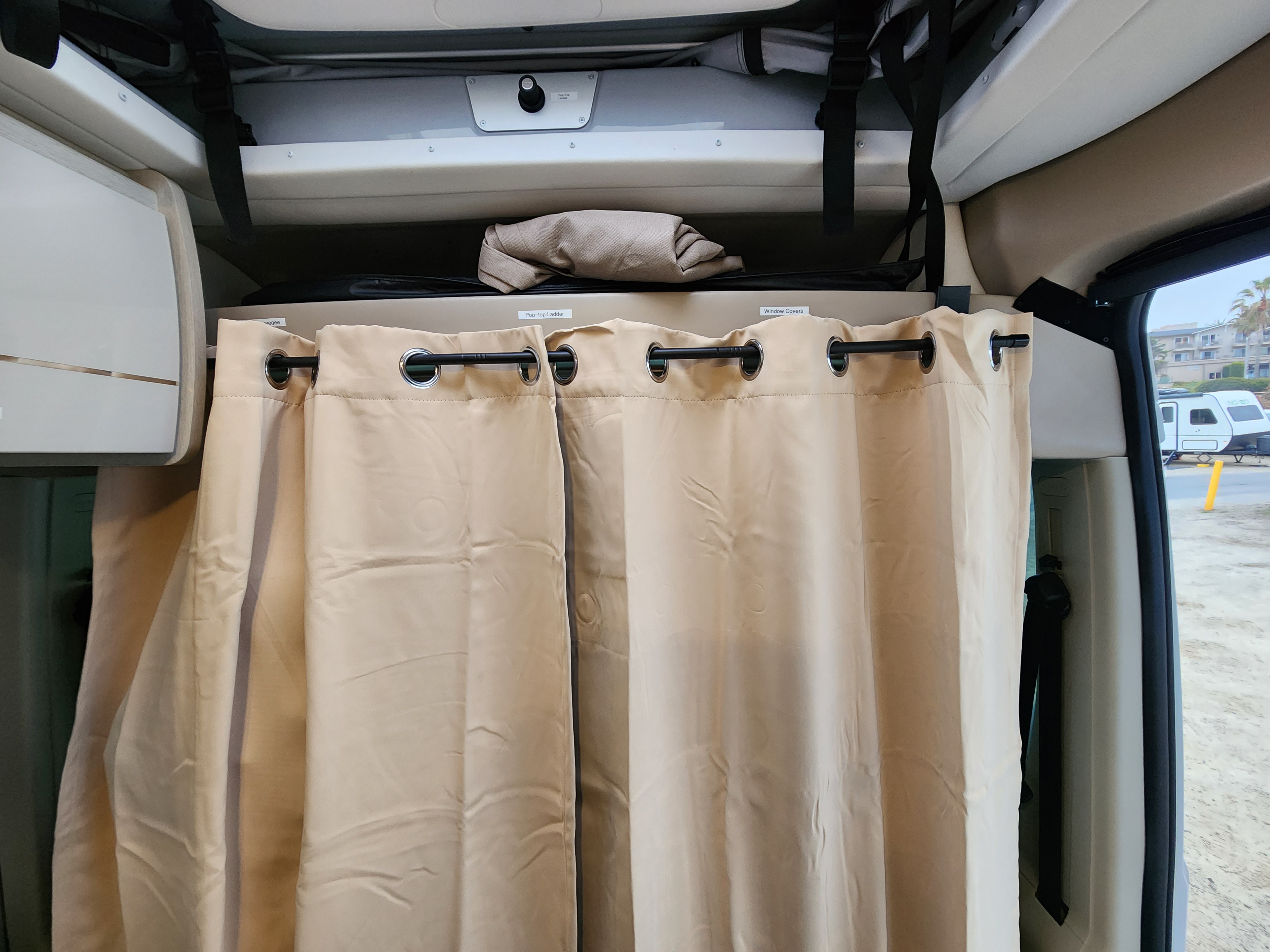 Inside an RV rental, featuring beige privacy curtains, overhead storage labeled 'Pop-top Ladder' & 'Window Covers'.. Thor Tellaro 2023