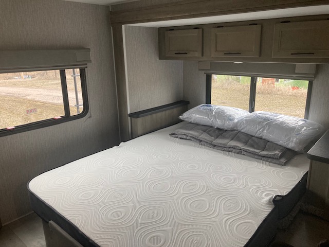 Cozy RV rental bedroom with a new mattress, pillows, and blanket on the bed. Windows look out to the outdoors.. Coachmen 26DS 2021