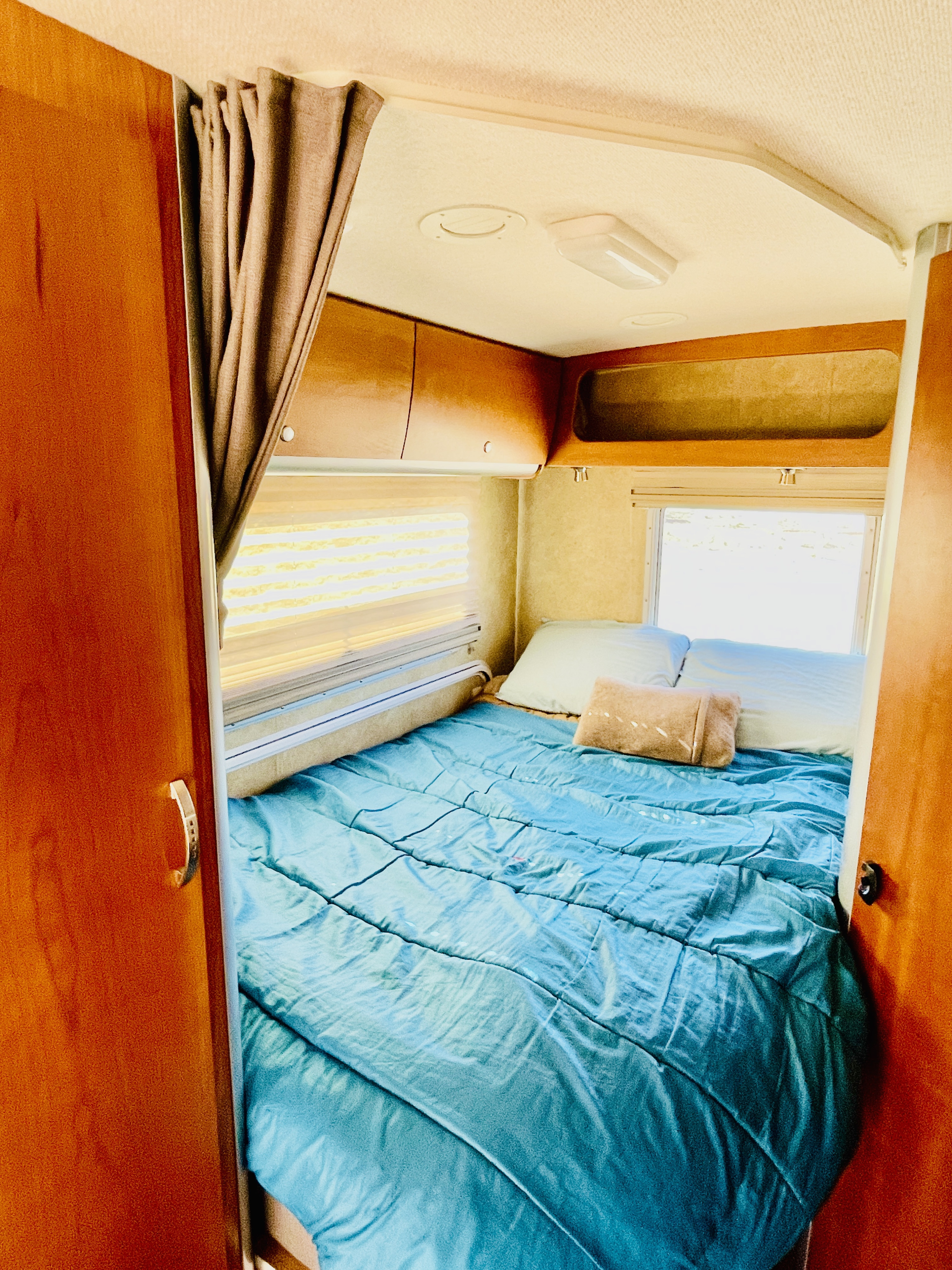 Cozy RV rental bedroom interior features a bed with a bright blue comforter, pillows, side windows, and overhead wooden cabinets.. Itasca Navion 2008