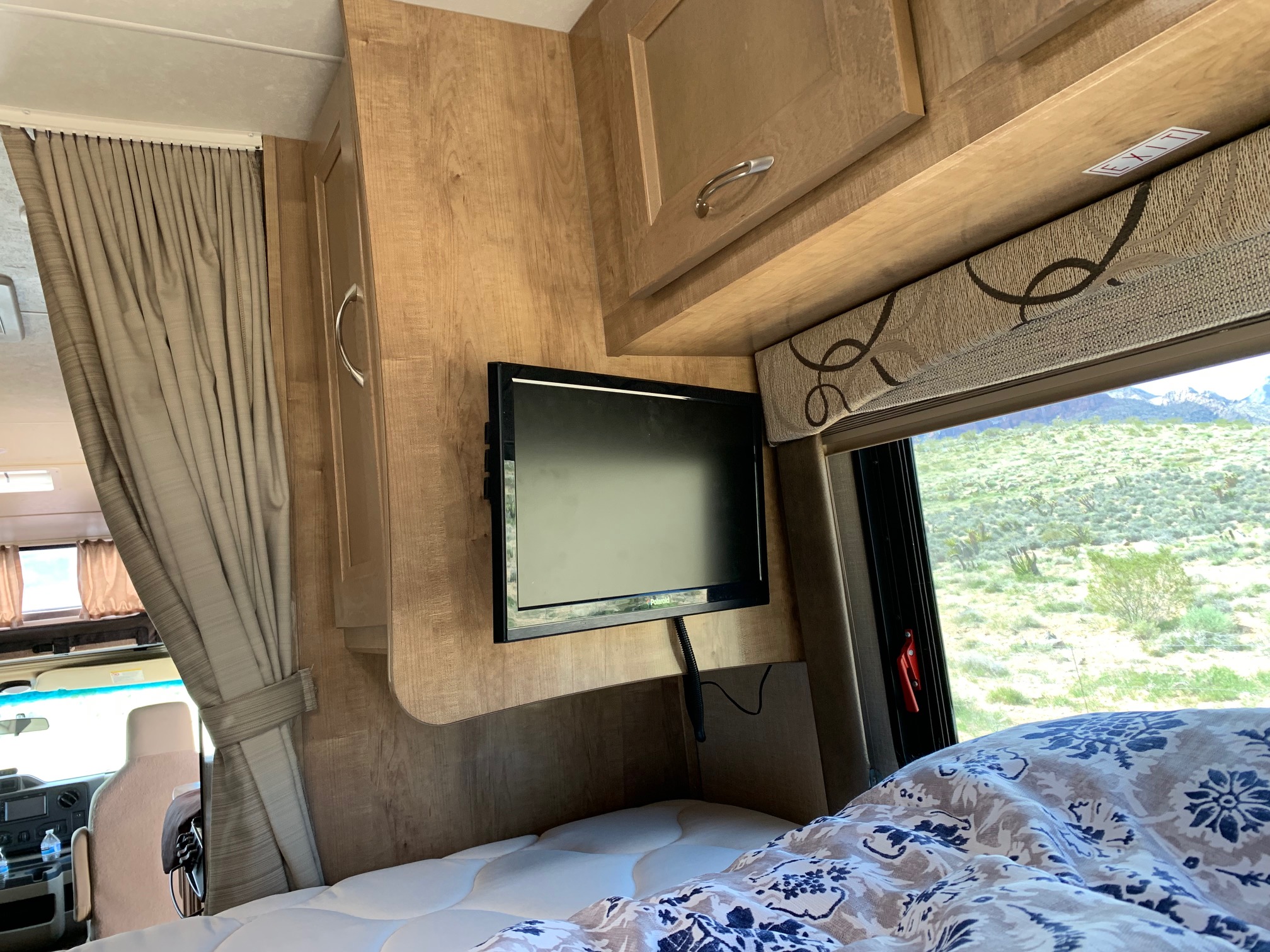 RV rental interior: bed with patterned duvet, mounted TV, wood cabinets, & desert views from window.. FORD E350 2020