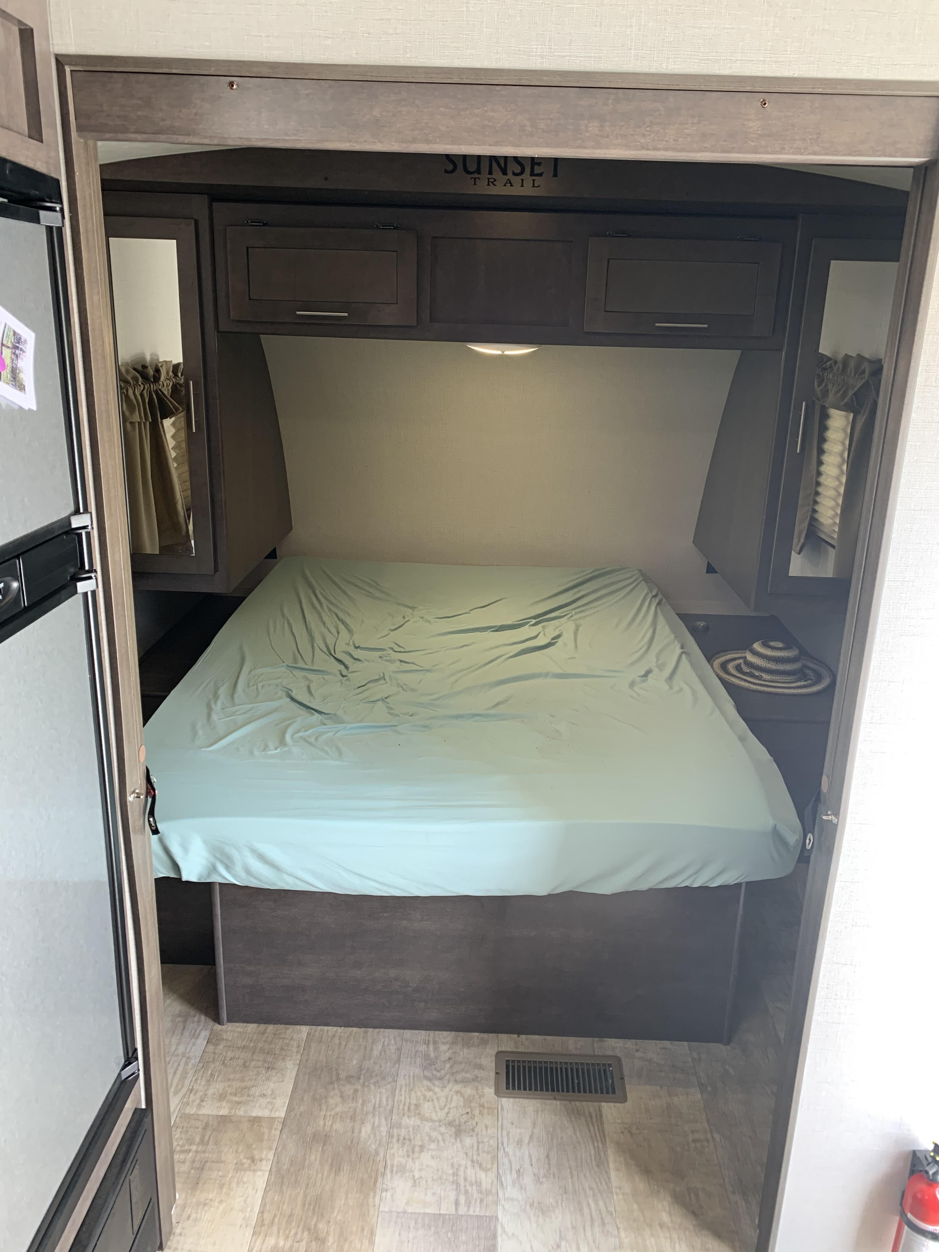 Cozy RV rental bedroom featuring an unmade bed, overhead cabinets, and a "Sunset Trail" logo. Refrigerator visible on left.. Crossroads Sunset Trail 2017