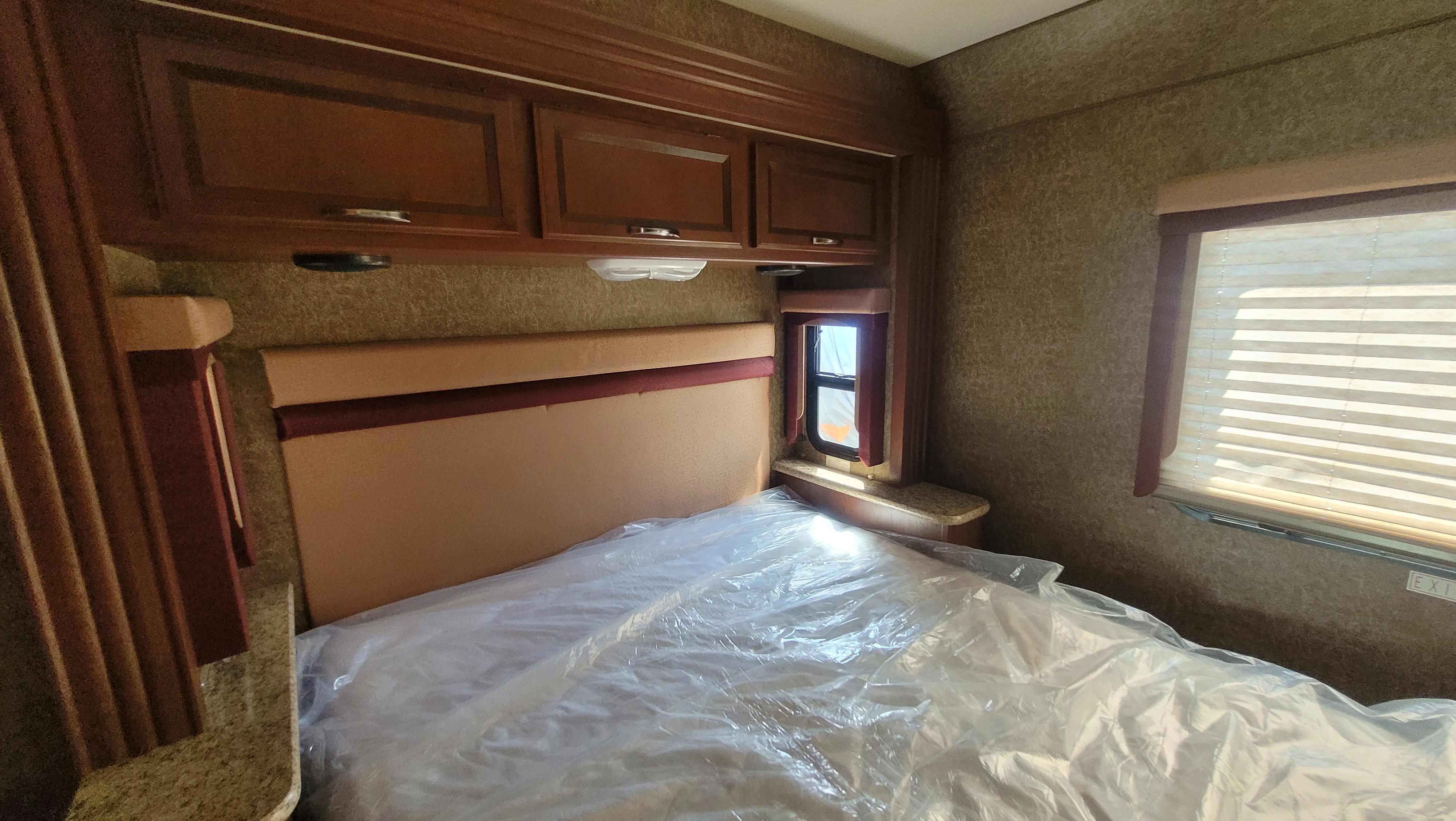Inside an RV rental: a comfortable bedroom with a bed covered in protective plastic, overhead cabinets, and a window with blinds.. A.C.E. A.C.E. Motorhome 2014
