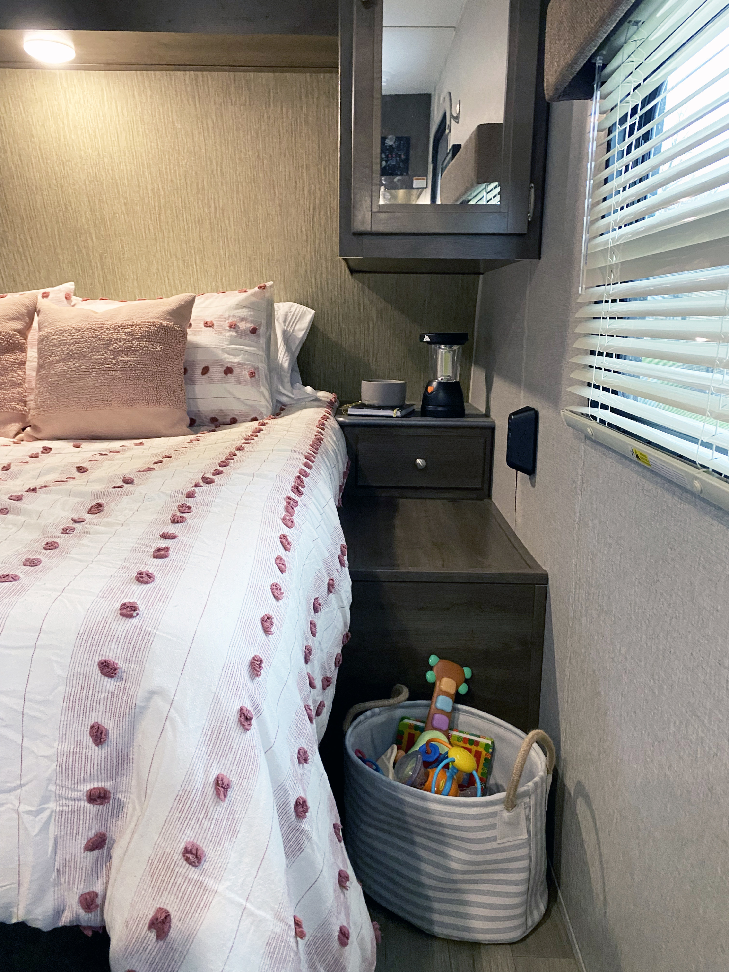 Cozy RV rental bedroom with pink-accented bed, practical nightstand with a lantern, and a striped toy basket. Ideal for family travel.. Dutchmen Coleman Lantern 2018