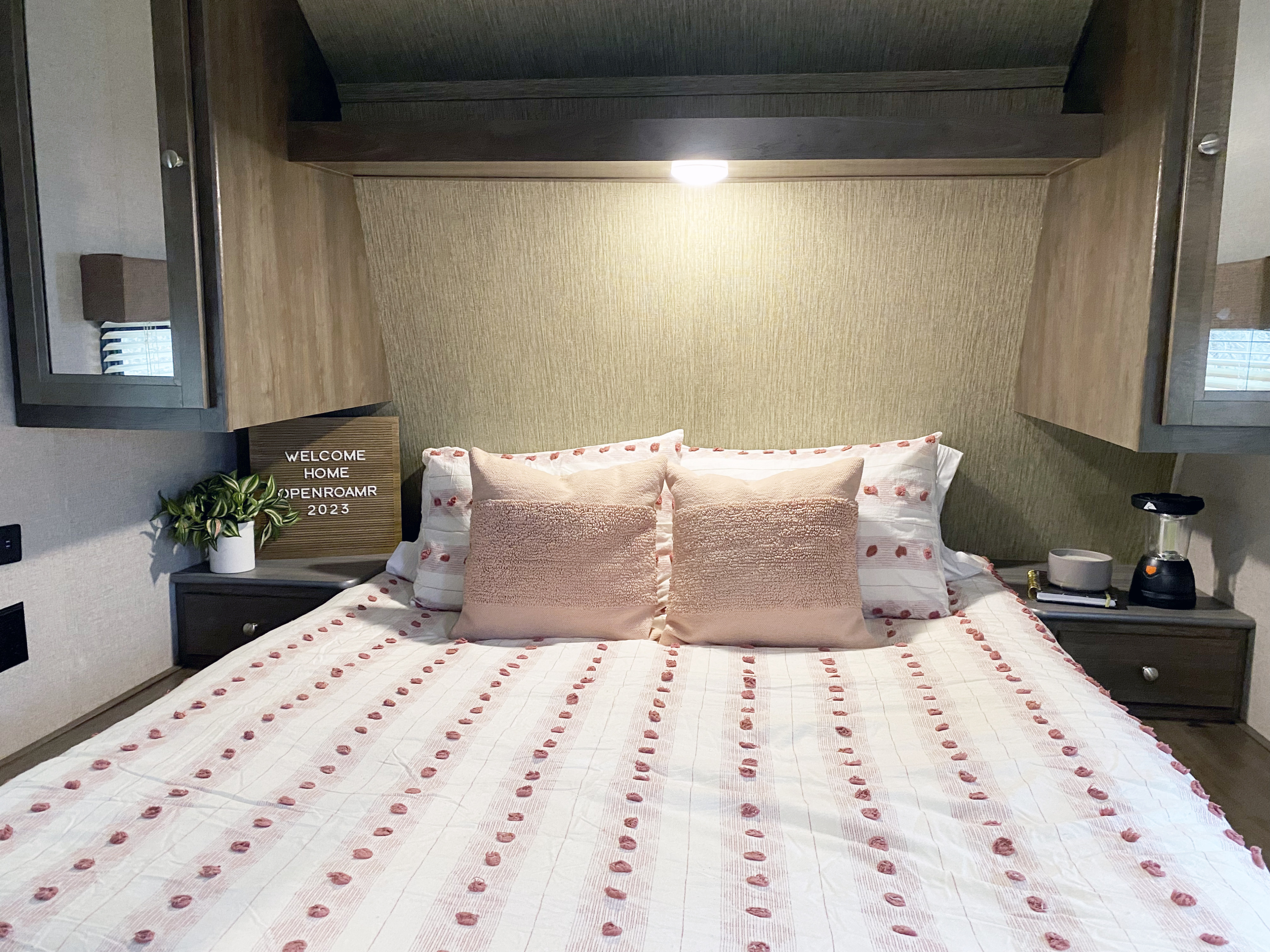 Cozy RV bedroom featuring pink and white bedding, a plant, and a 'Welcome Home' sign, ideal for your next rv rental.. Dutchmen Coleman Lantern 2018