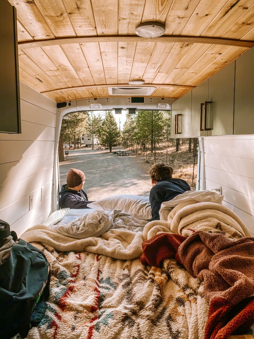 Cozy rv rental interior: two people on a soft bed looking out at a tranquil, tree-lined campground. Perfect for travel.. Ford Custom Transit 2019