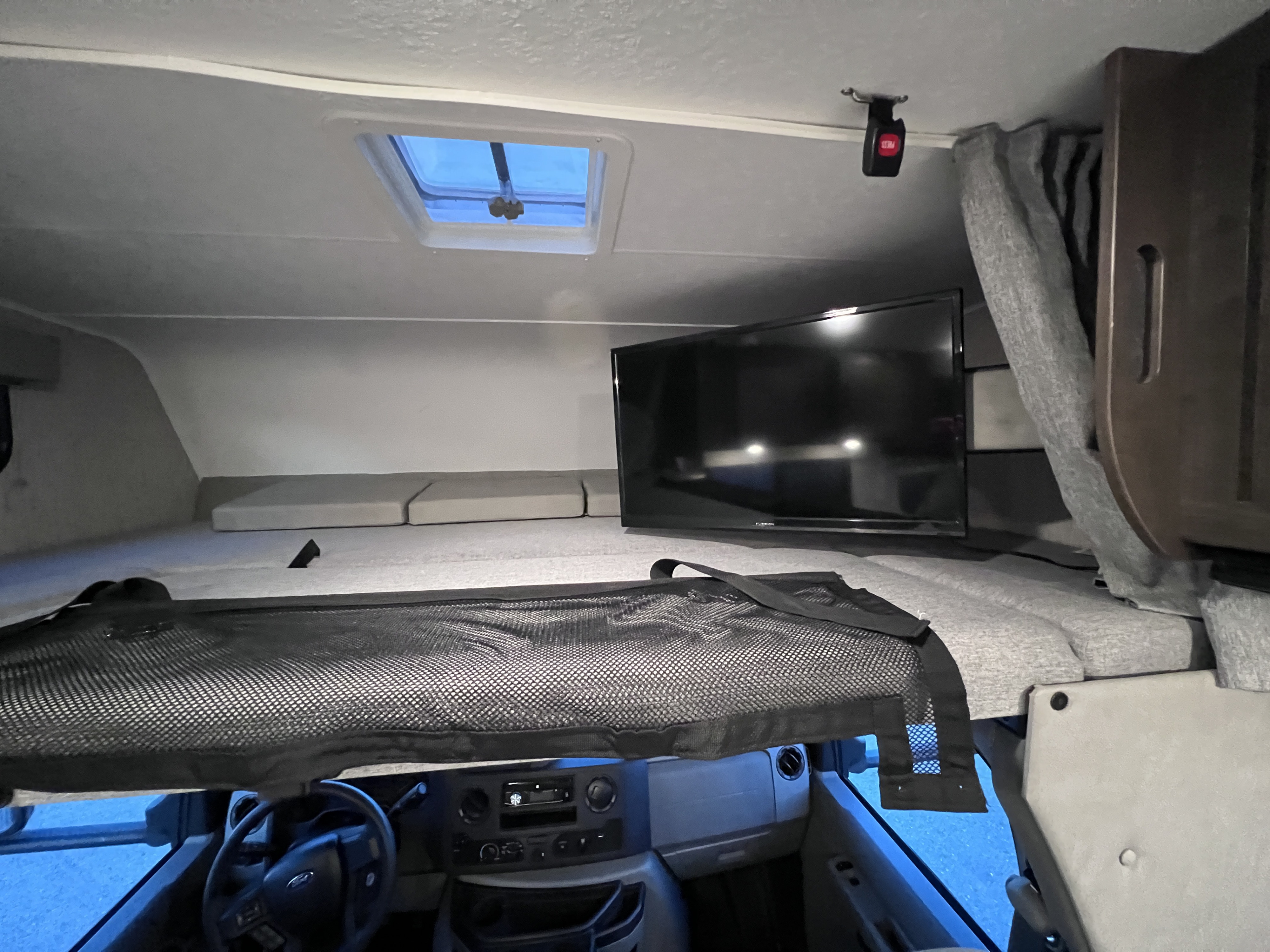 Inside an RV rental: view of the overhead bunk bed with cushions, TV, ceiling skylight, and a safety net above the cab.. Forest River Coachmen Leprechaun 2022
