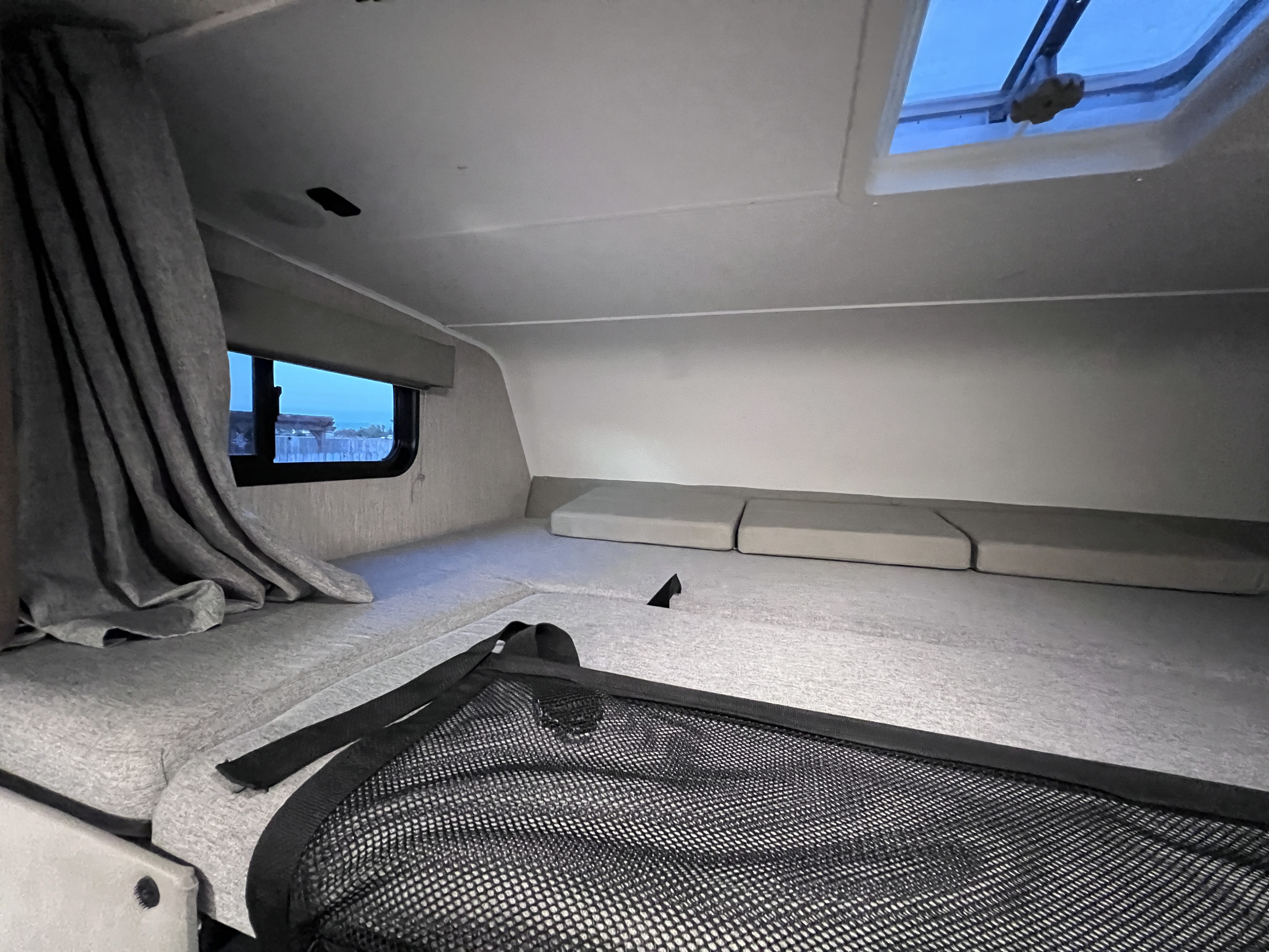 An rv rental interior featuring a cab-over bed with gray cushions, a window looking outside, and a ceiling skylight.. Forest River Coachmen Leprechaun 2022