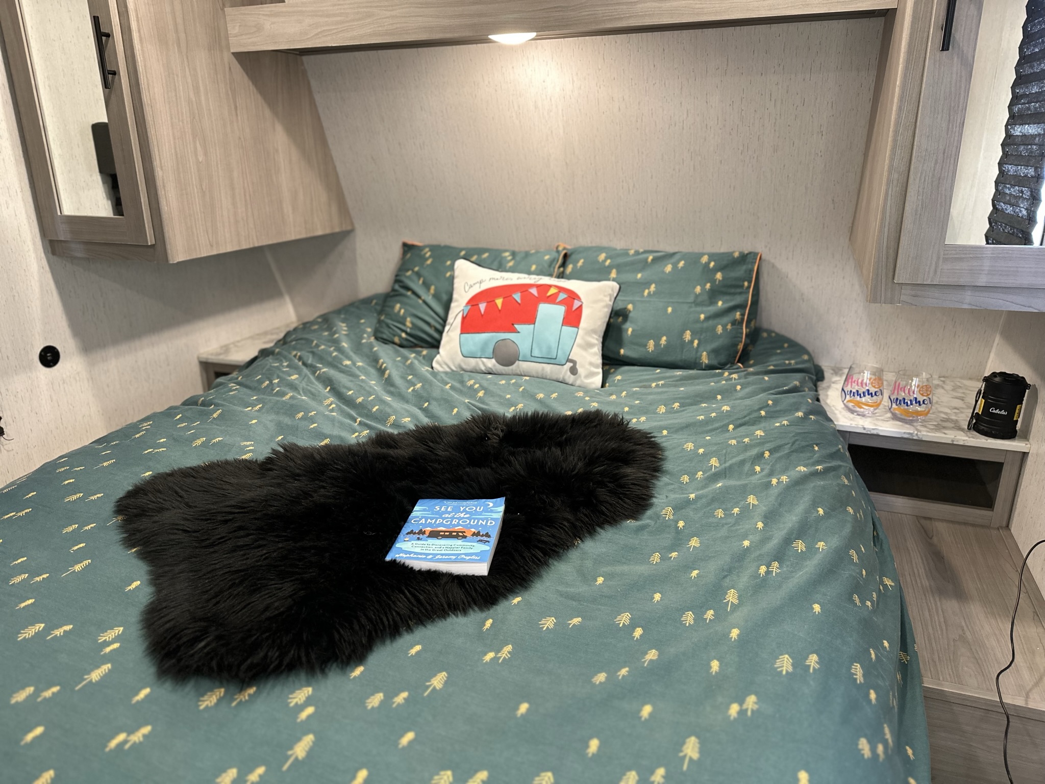 Inside a cozy RV rental: teal bed with tree print, camper pillow, and a camping book. Ideal for travel adventures.. Dutchmen Coleman 2022