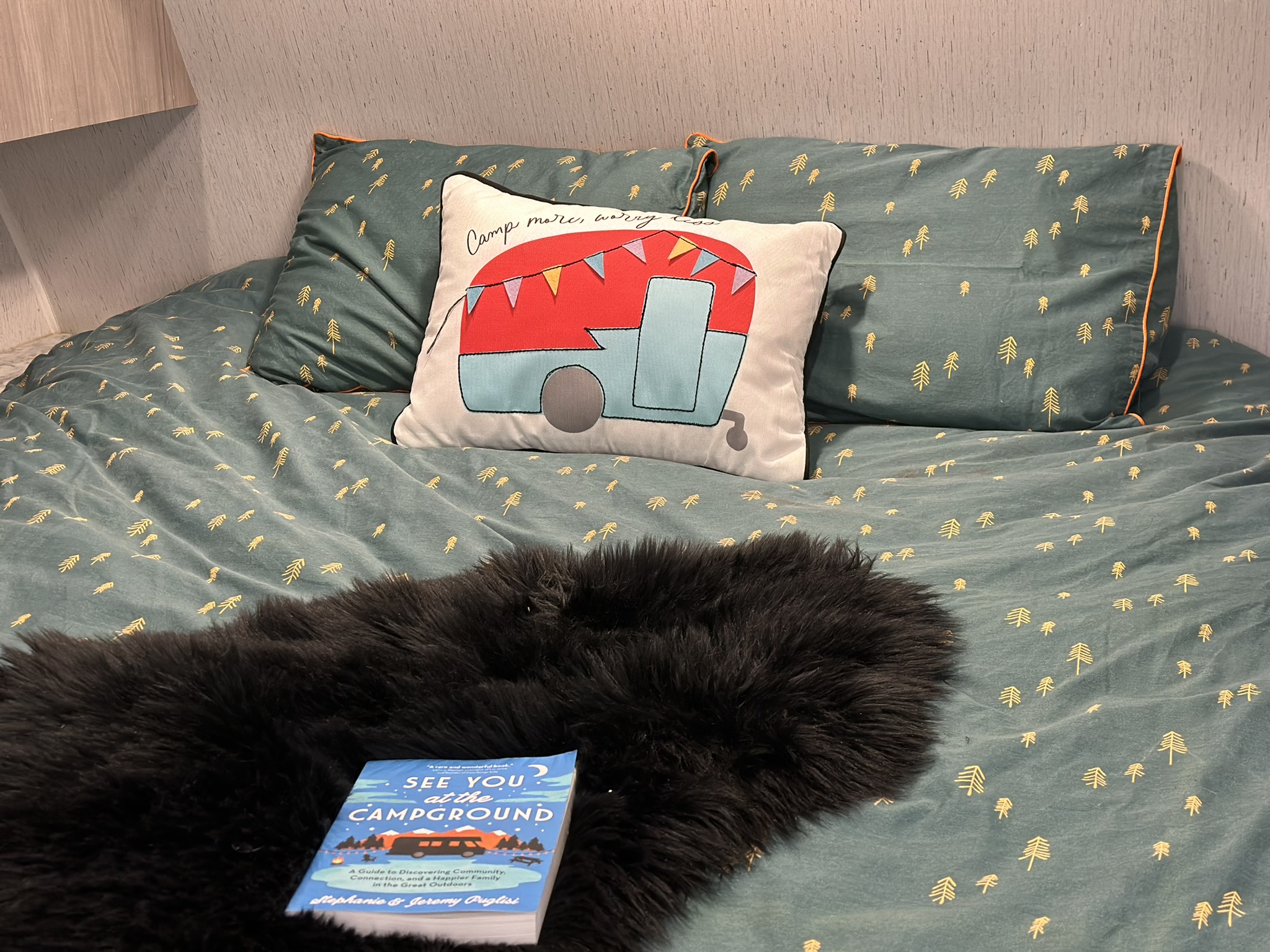 Cozy RV rental bed with charming tree-patterned bedding, a camper pillow, and a 'See You at the Campground' book.. Dutchmen Coleman 2022