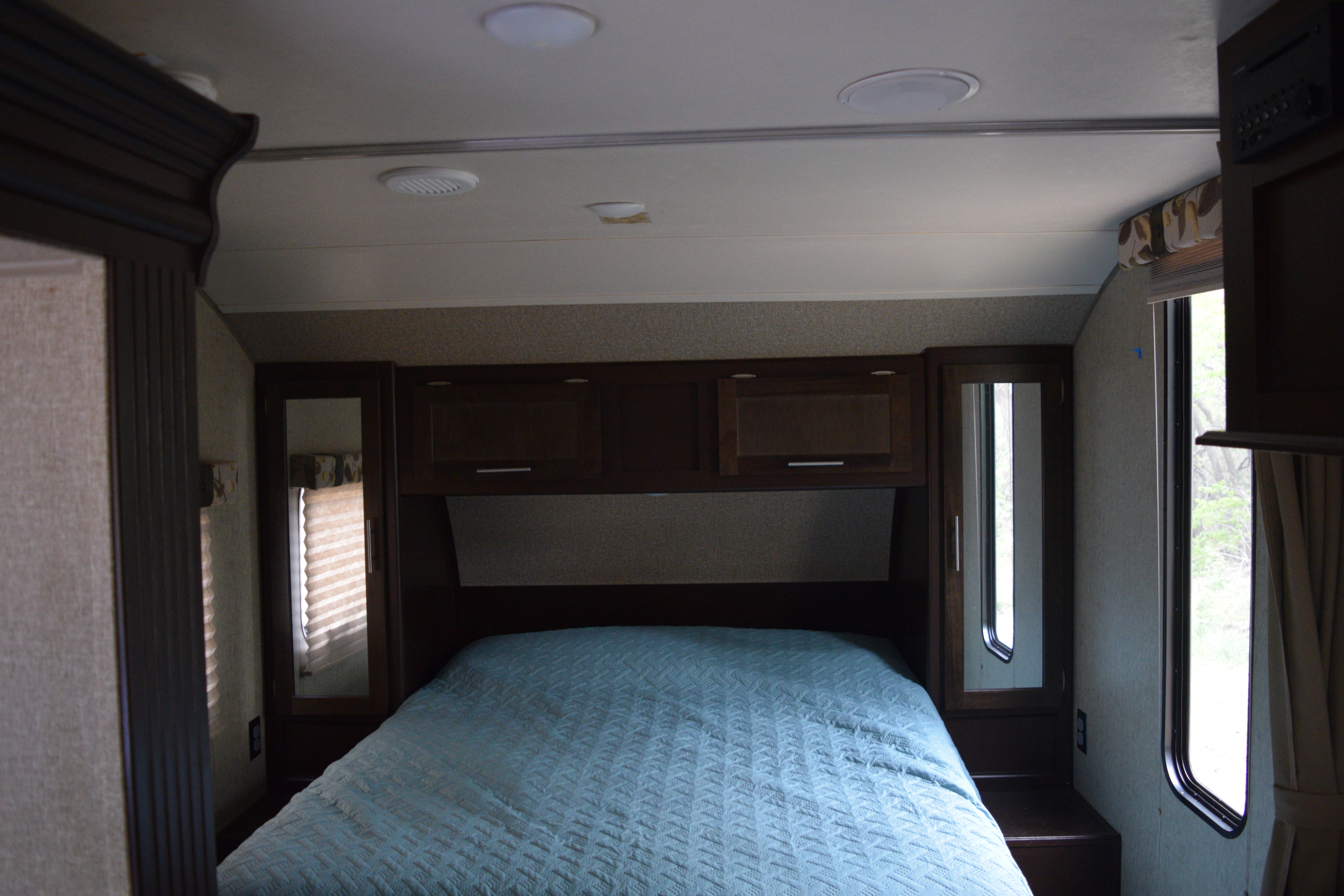 Cozy RV rental bedroom featuring a blue quilted bed, dark wood cabinets, and windows showing outdoor trees.. Forest River Cherokee 2017