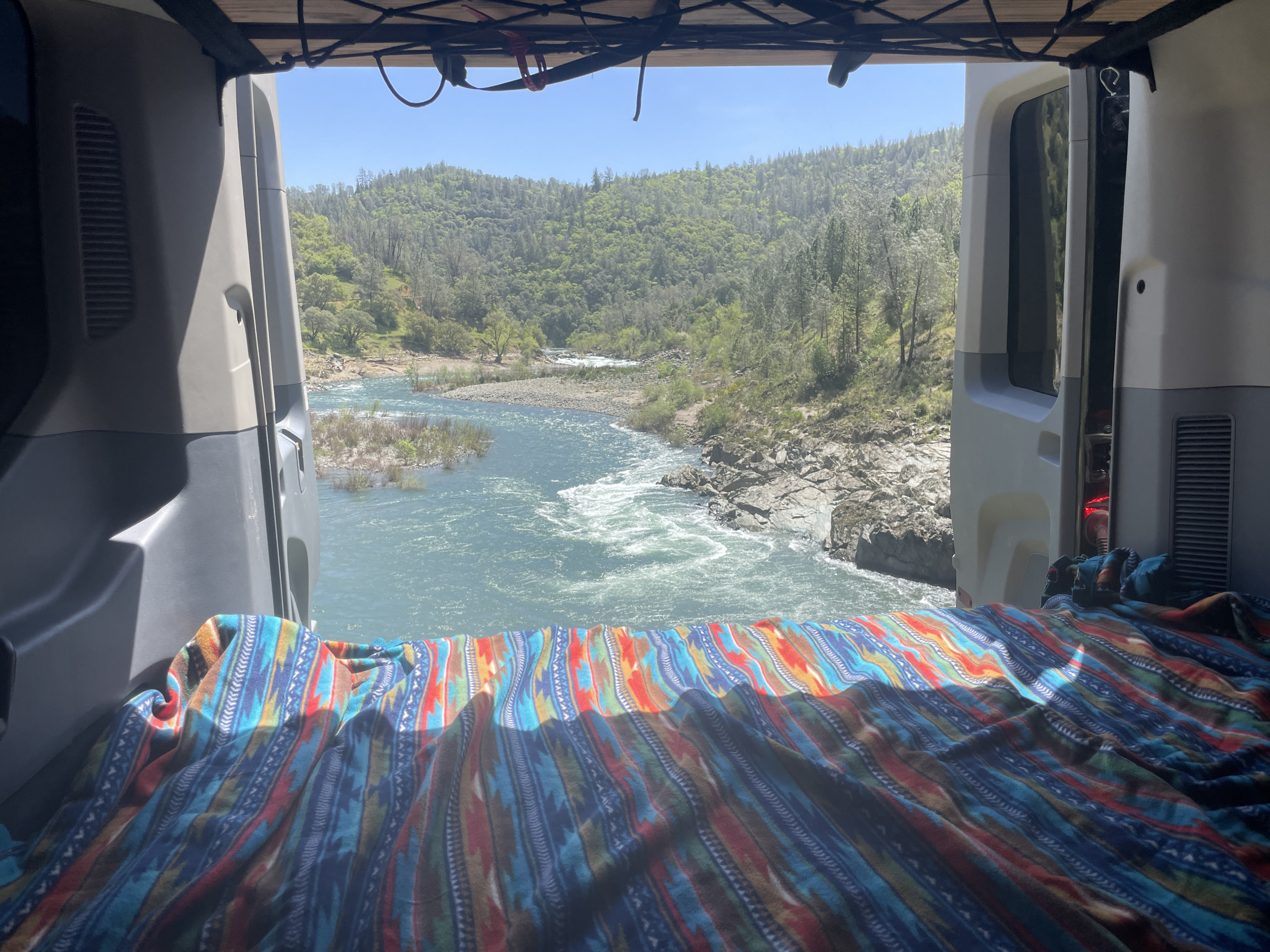 Scenic view from an **rv rental** van overlooking a vibrant river and lush mountains. A colorful blanket covers the bed, perfect for **van life**.. Ford Transit 2017