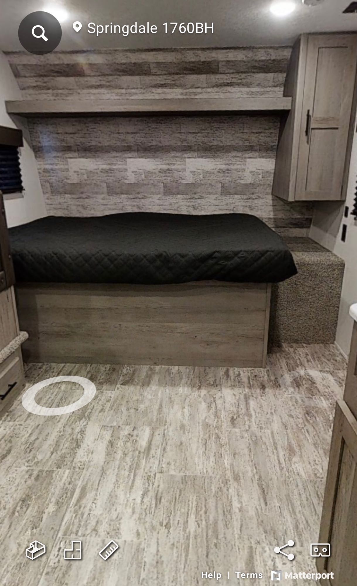 An RV rental interior features a cozy bedroom area with a dark quilted bed, light wood-look paneling, and plank flooring.. Keystone Springdale 2022