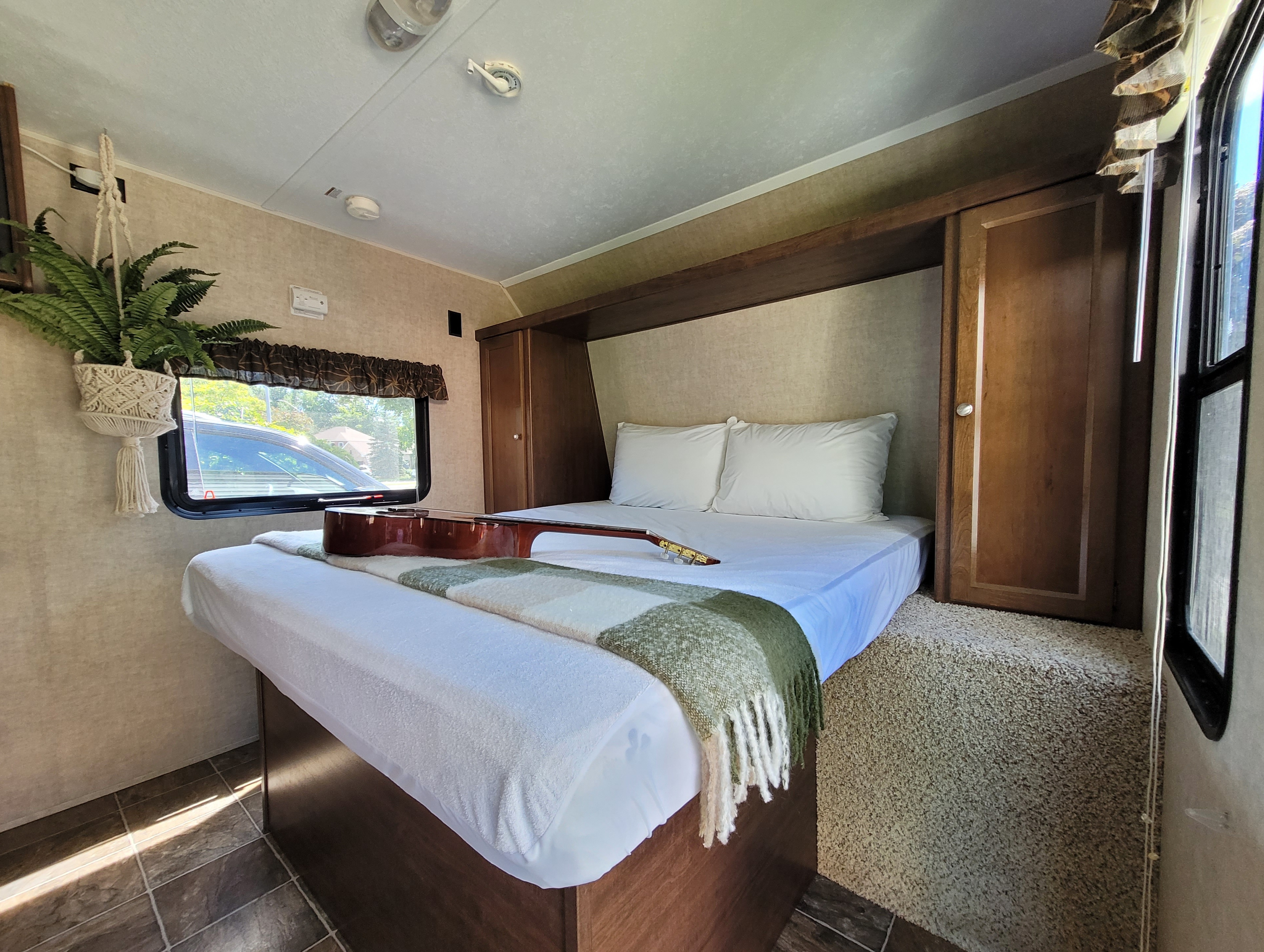 Spacious RV rental bedroom with a cozy bed, acoustic guitar, and hanging fern. Features warm wood cabinetry and natural light.. Keystone RV Hideout LHS 2016