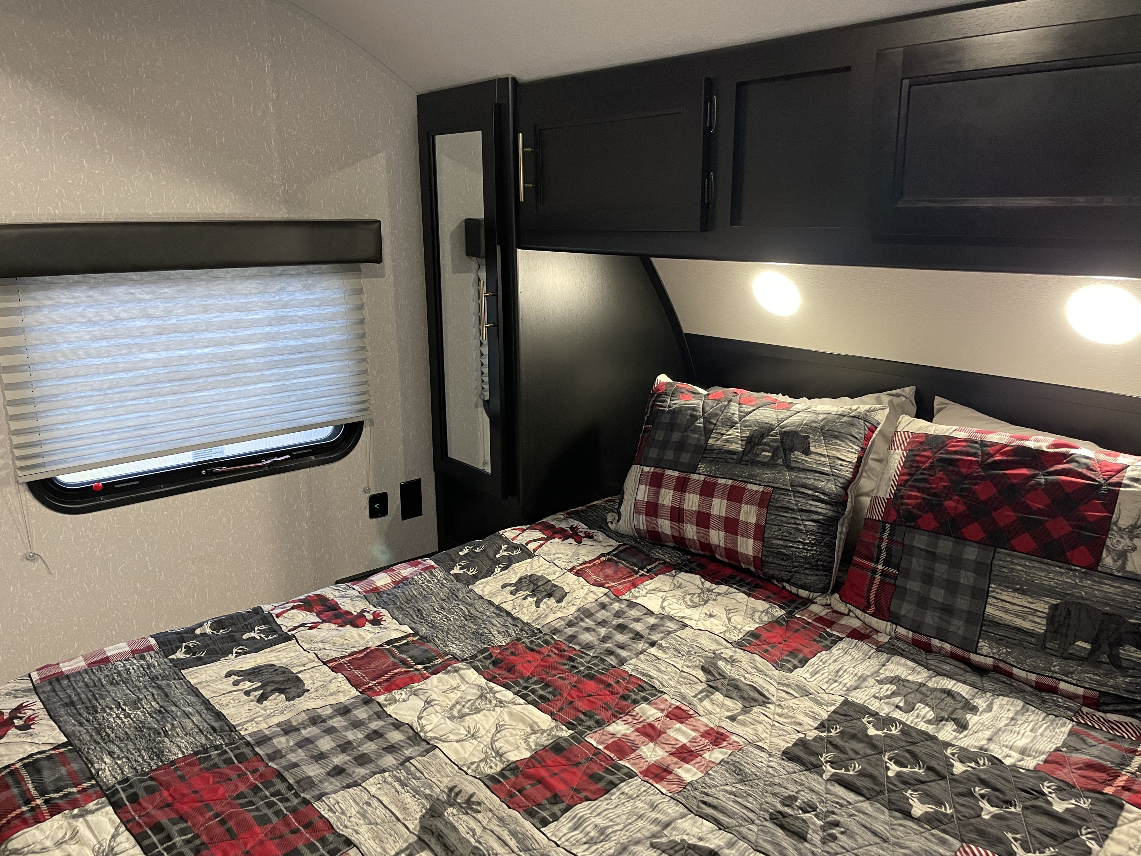 Cozy RV rental bedroom features a plaid & animal-patterned quilt on the bed, dark cabinets, and a window with blinds.. Forest River Cherokee Grey Wolf 2022