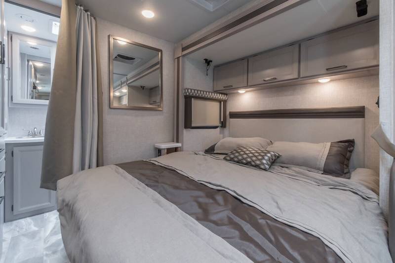 Modern RV rental bedroom featuring a comfortable bed, overhead storage, and an adjacent compact bathroom with vanity.. Thor Delano 2023
