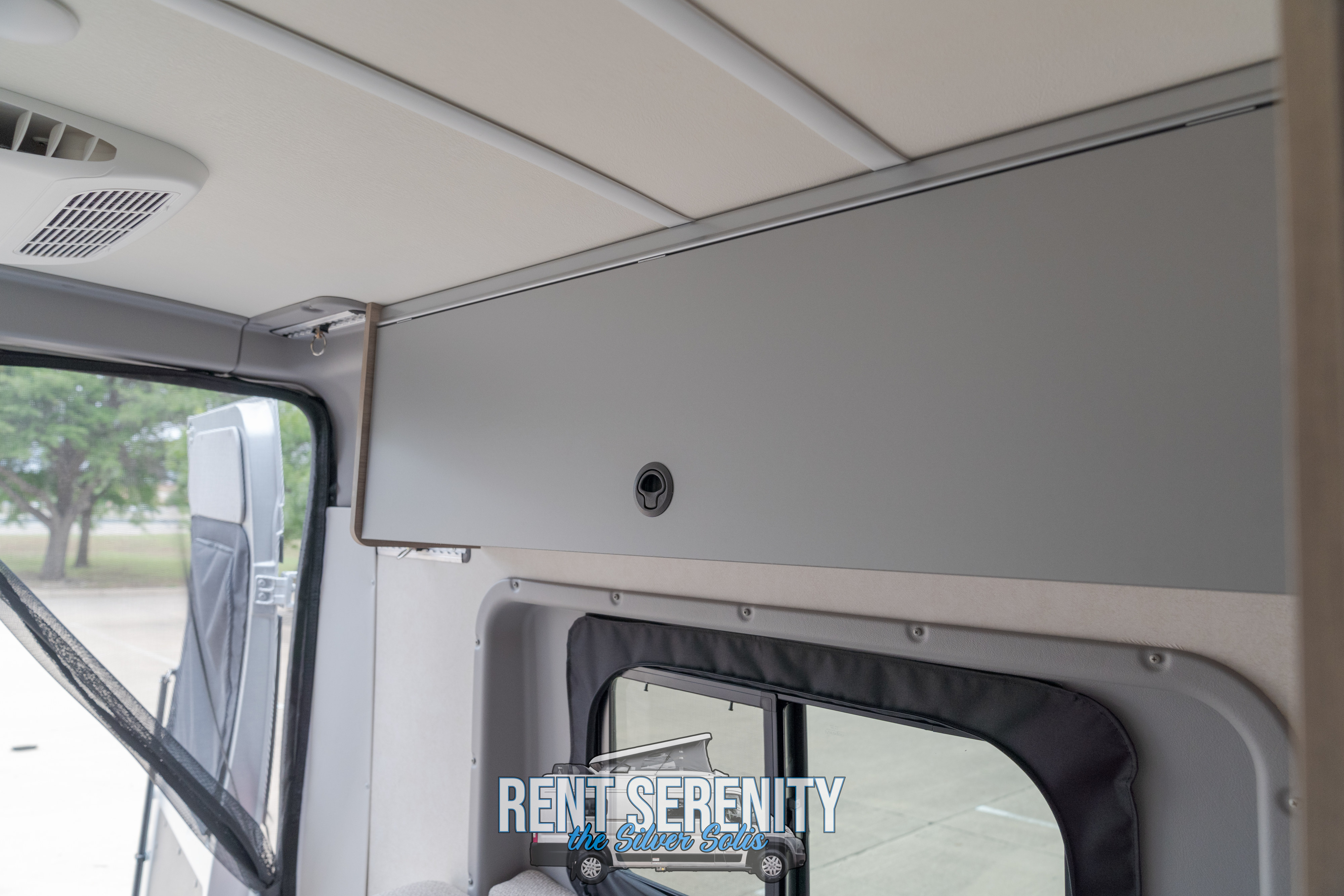 Inside a comfortable RV rental: A beige ceiling, AC vent, grey overhead cabinet, and a window with a bug screen.. Winnebago Solis 2023