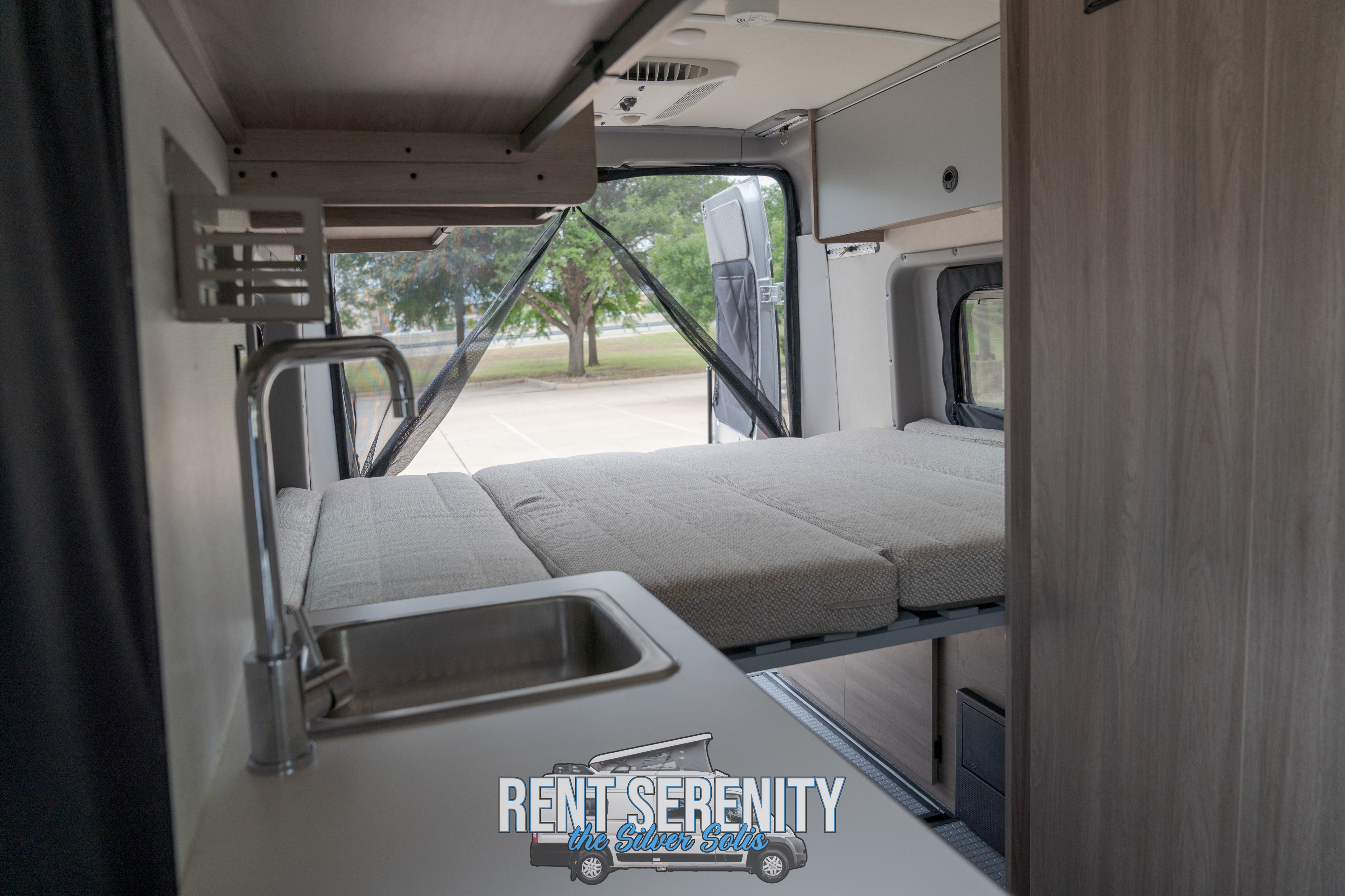 RV rental: Camper van interior with a made bed, kitchen sink, and open rear doors with a mesh screen, overlooking a parking lot.. Winnebago Solis 2023