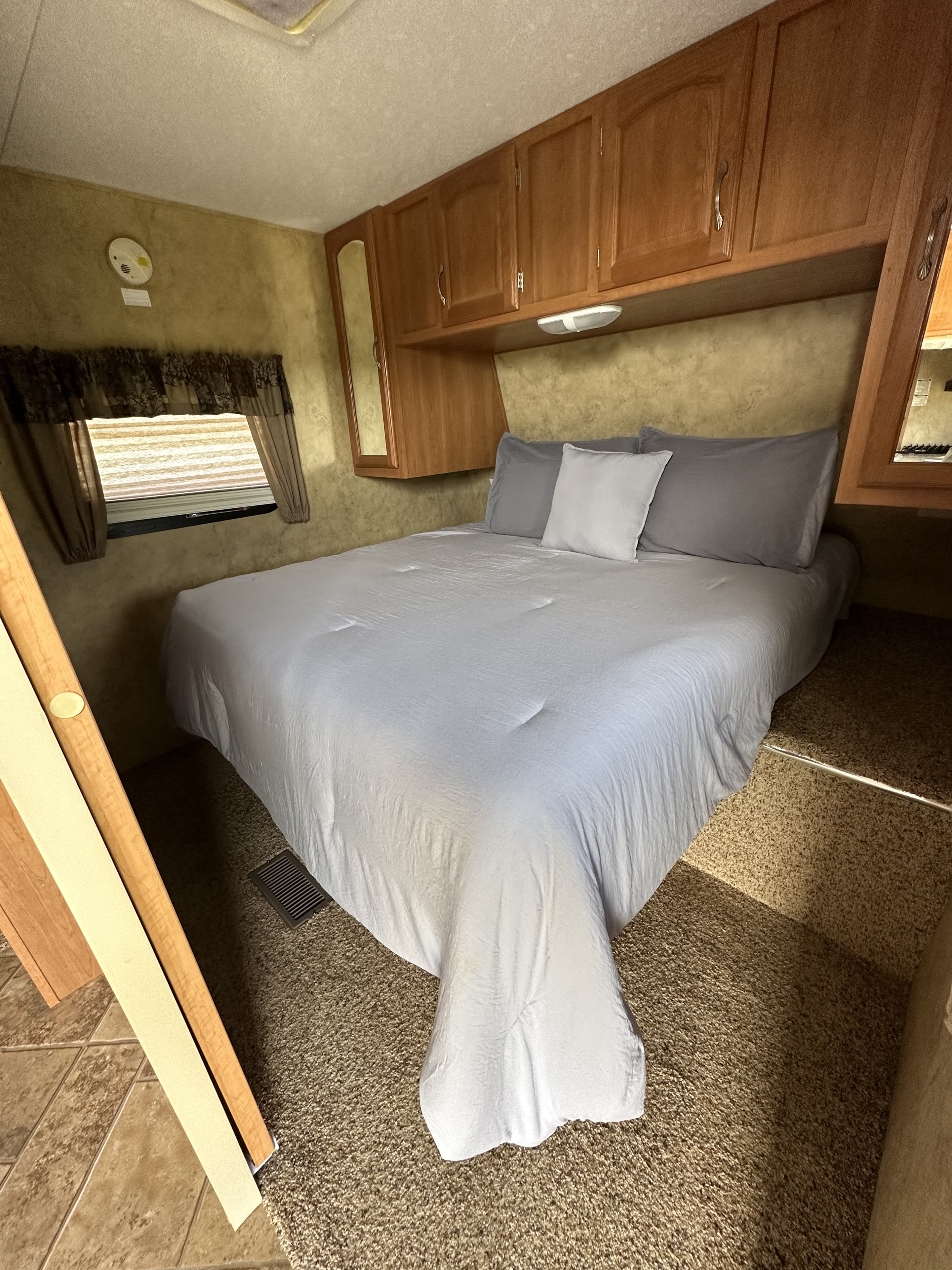 Cozy RV rental bedroom featuring a spacious bed with light blue bedding, overhead cabinets, and window views.. Keystone RV Cougar X-Lite 2009
