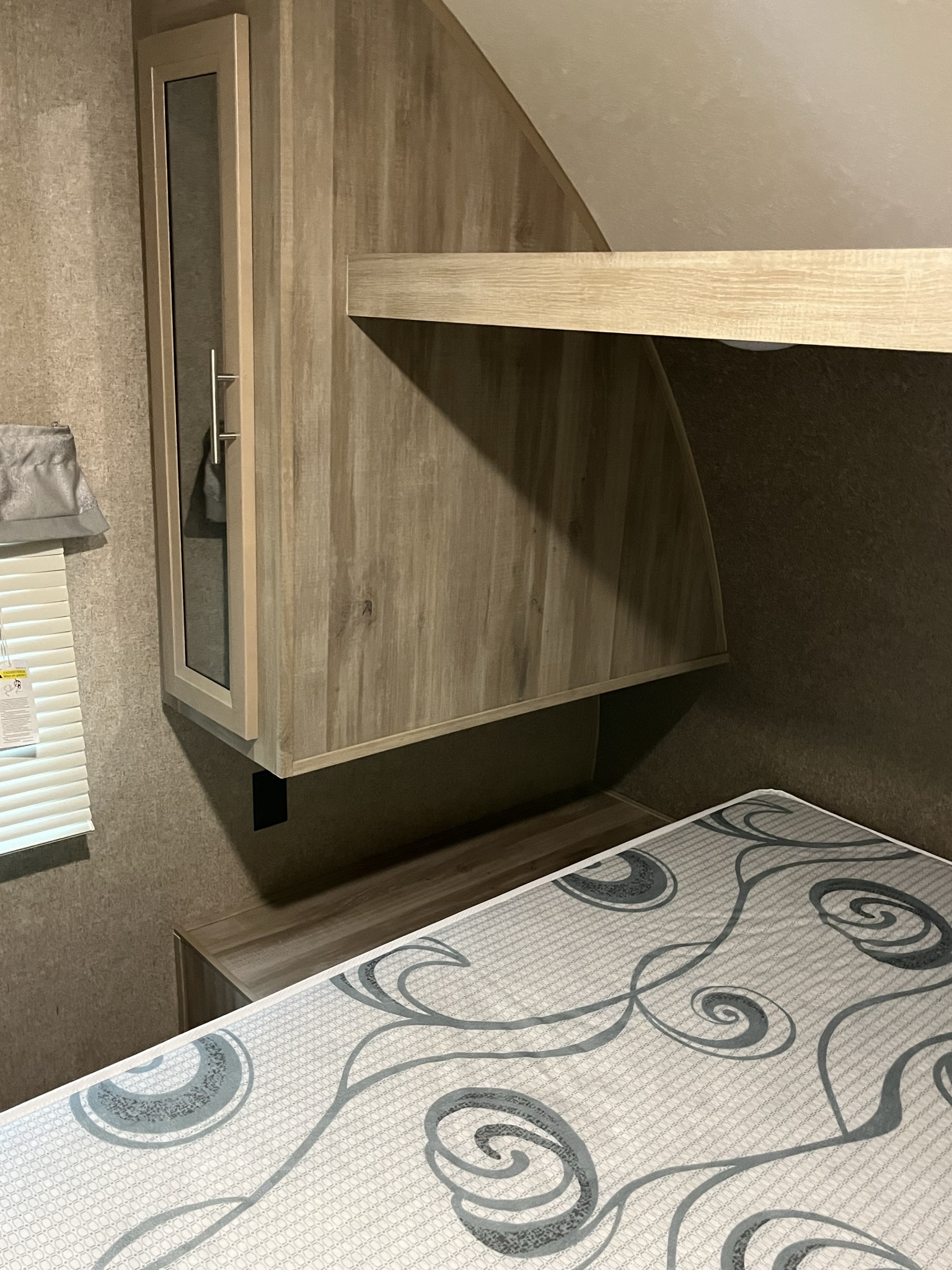 RV rental interior: a bed with a patterned mattress, an overhead wooden cabinet with a glass door, and a shelf.. Forest River Coachmen Catalina SBX 2019