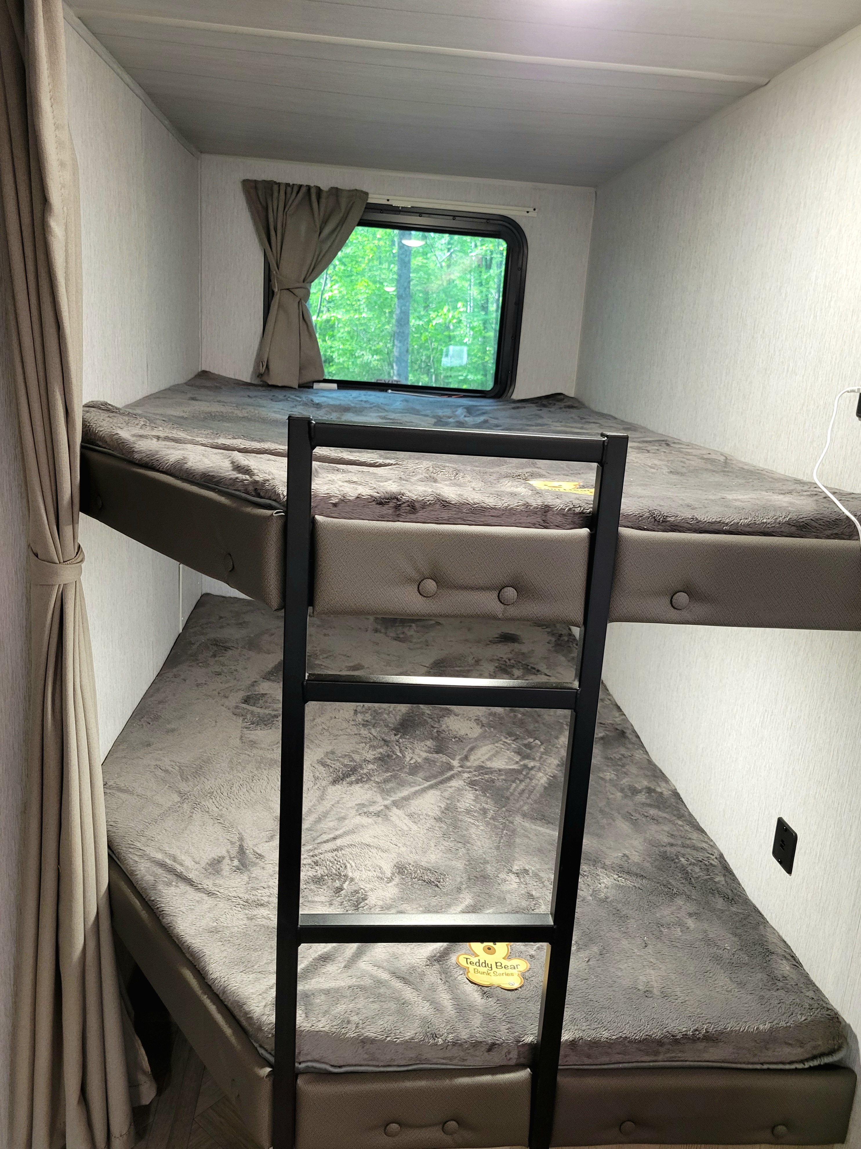 Cozy bunk beds in an RV rental, featuring soft gray blankets and a "Teddy Bear" series mattress, with a nature view from the window.. Forest River 31bhxl 2023