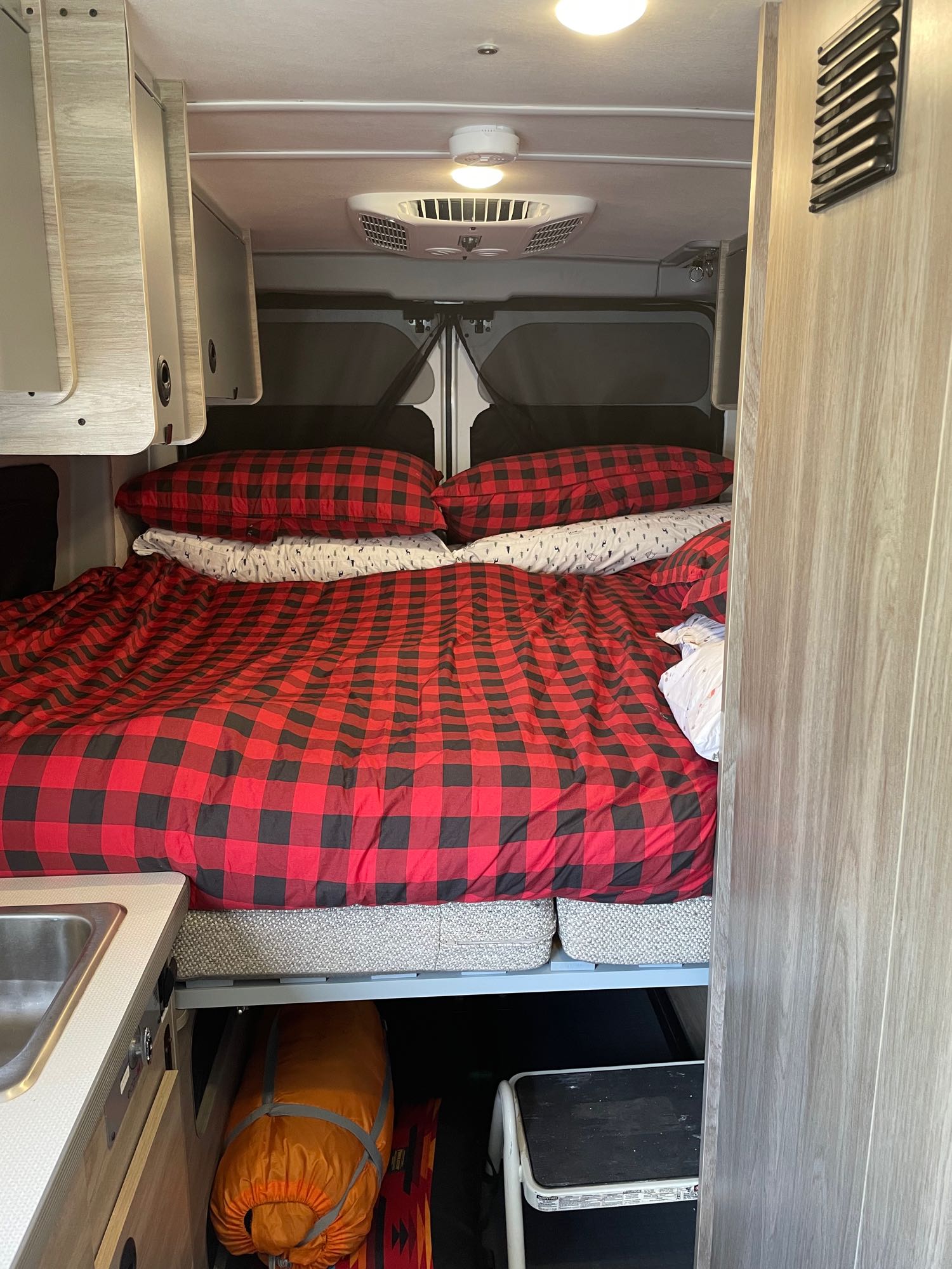 Inside an RV rental: a cozy bed with red plaid bedding, overhead cabinets, and under-bed storage for travel gear.. Winnebago Winnebago 2023