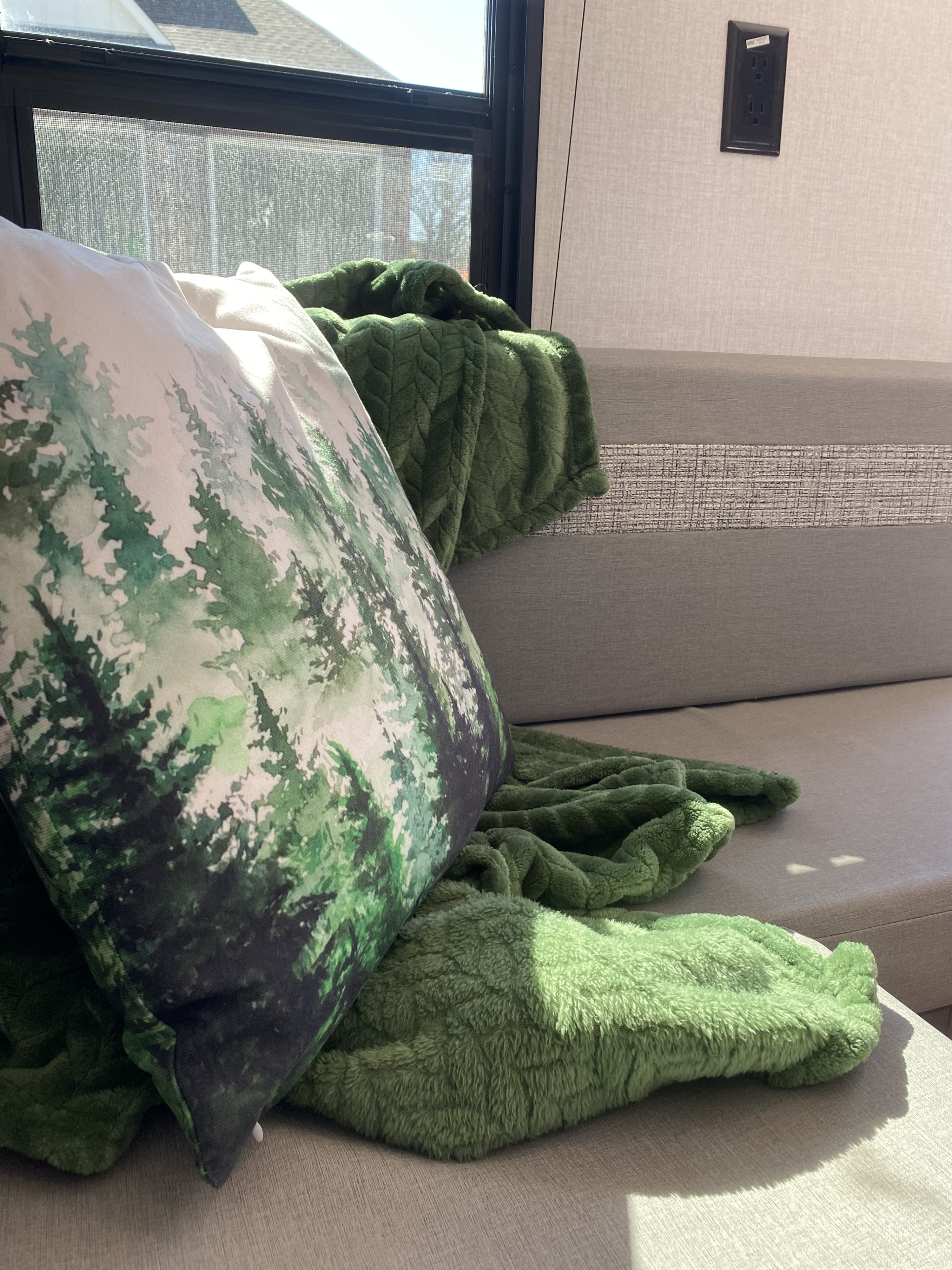 Enjoy the comfort of your RV rental: a plush green blanket, forest-print pillow, and sunlit sofa. Perfect for relaxation.. Forest River Coachmen Clipper 182DBU 2022