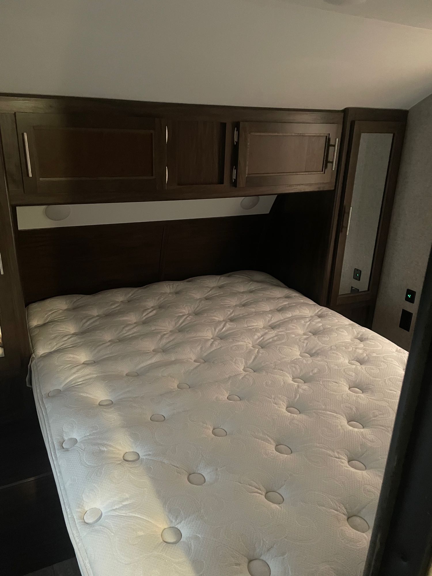 Inside this RV rental: A compact bedroom with a pristine tufted mattress, dark wood overhead cabinets, and a side mirror.. Forest River Cherokee Grey Wolf 2019