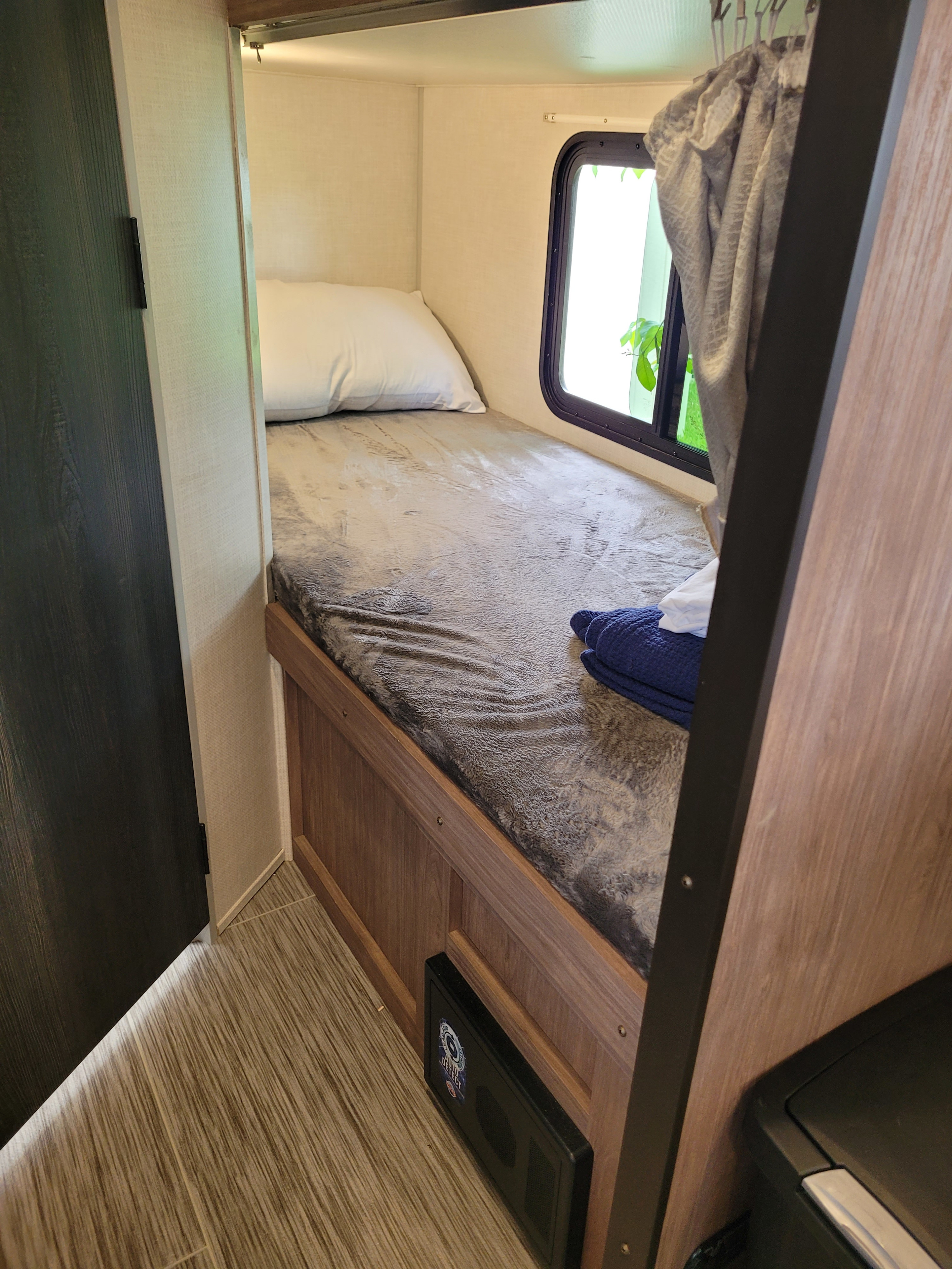 Inside an RV rental: a cozy compact bed with a soft blanket, white pillow, window view, and blue towels.. Forest River No Boundaries 2022