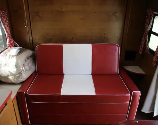 A stylish red and white convertible sofa in a vintage RV rental interior, with warm wood paneling and stored bedding.. Gulf Stream Vintage Cruiser 2021