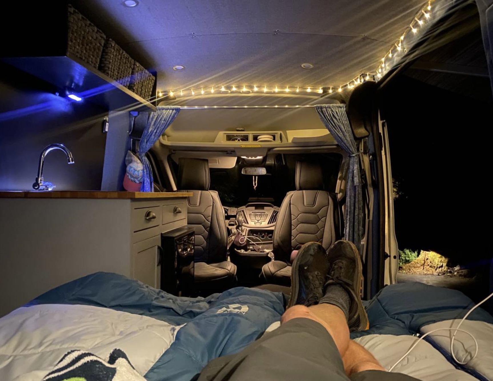 Enjoying van life in a comfortable RV rental at night. Interior features a bed, kitchenette, and bright string lights.. Ford Transit 2019