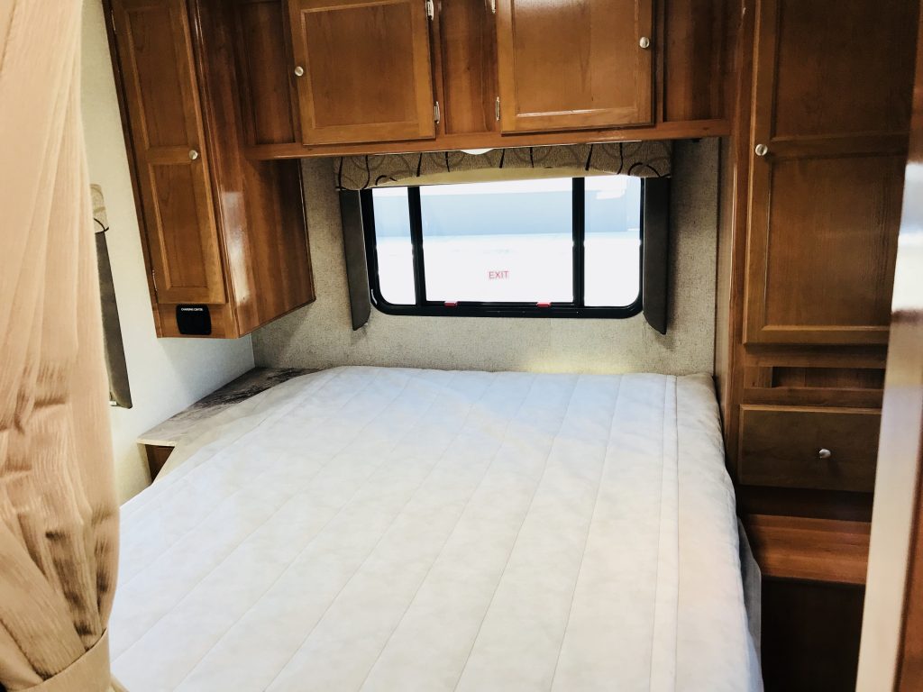 RV rental bedroom: a compact interior features a white bed, window with exit sign, and wood cabinetry.. Coachmen Leprechaun 2016