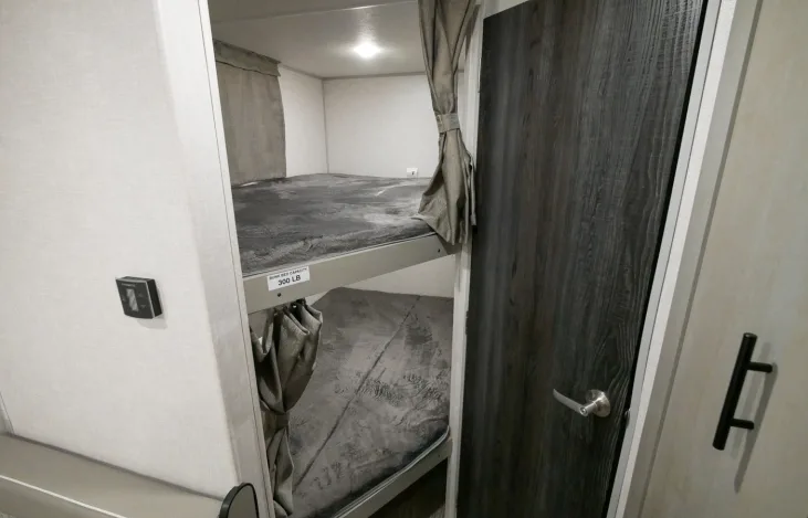 RV rental bunk beds with soft gray bedding & tied-back privacy curtains. Features a dark wood door & climate control.. Forest River IBEX 19MBH 2022