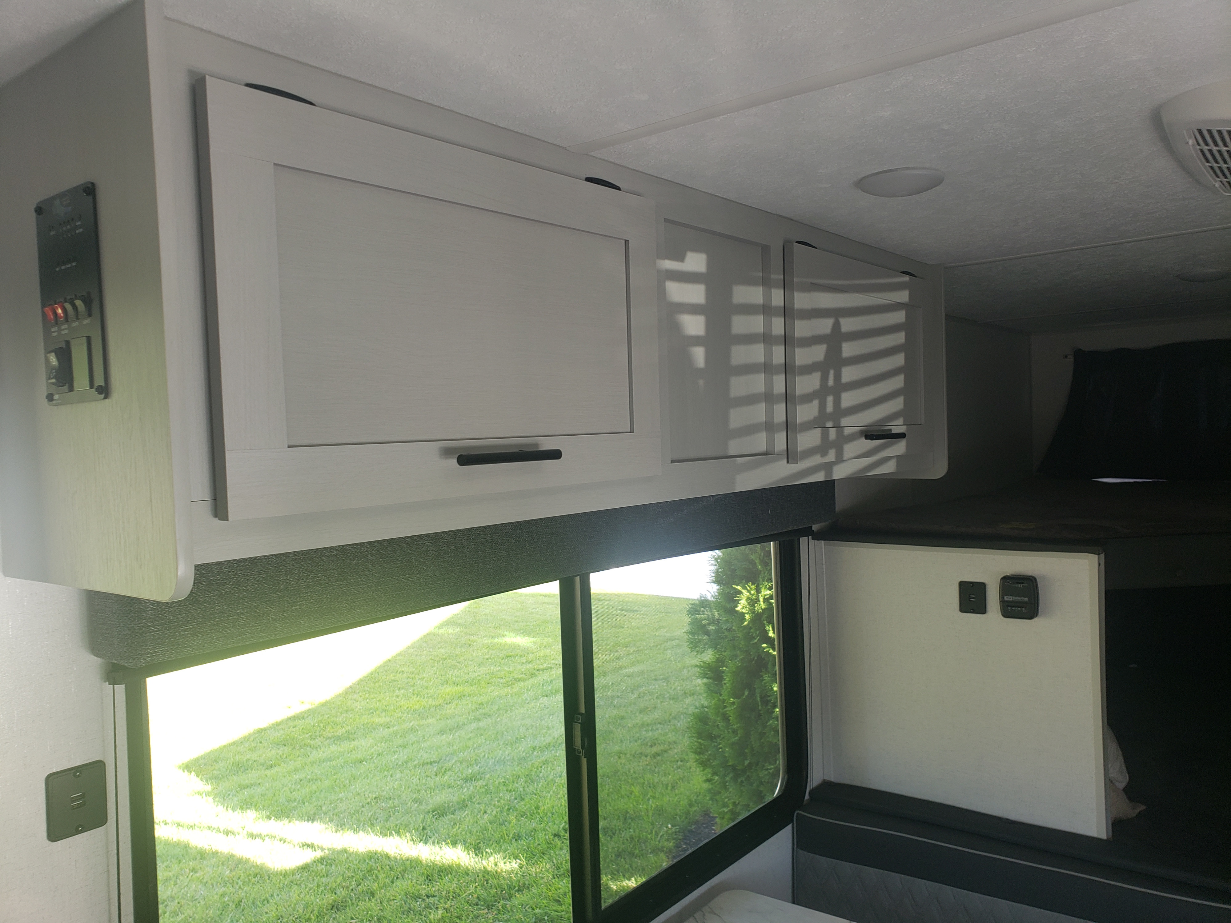 RV rental interior: Light overhead cabinets, a large window with a green outdoor view, system controls, and a bunk bed.. Forest River Wildwood FSX 2022