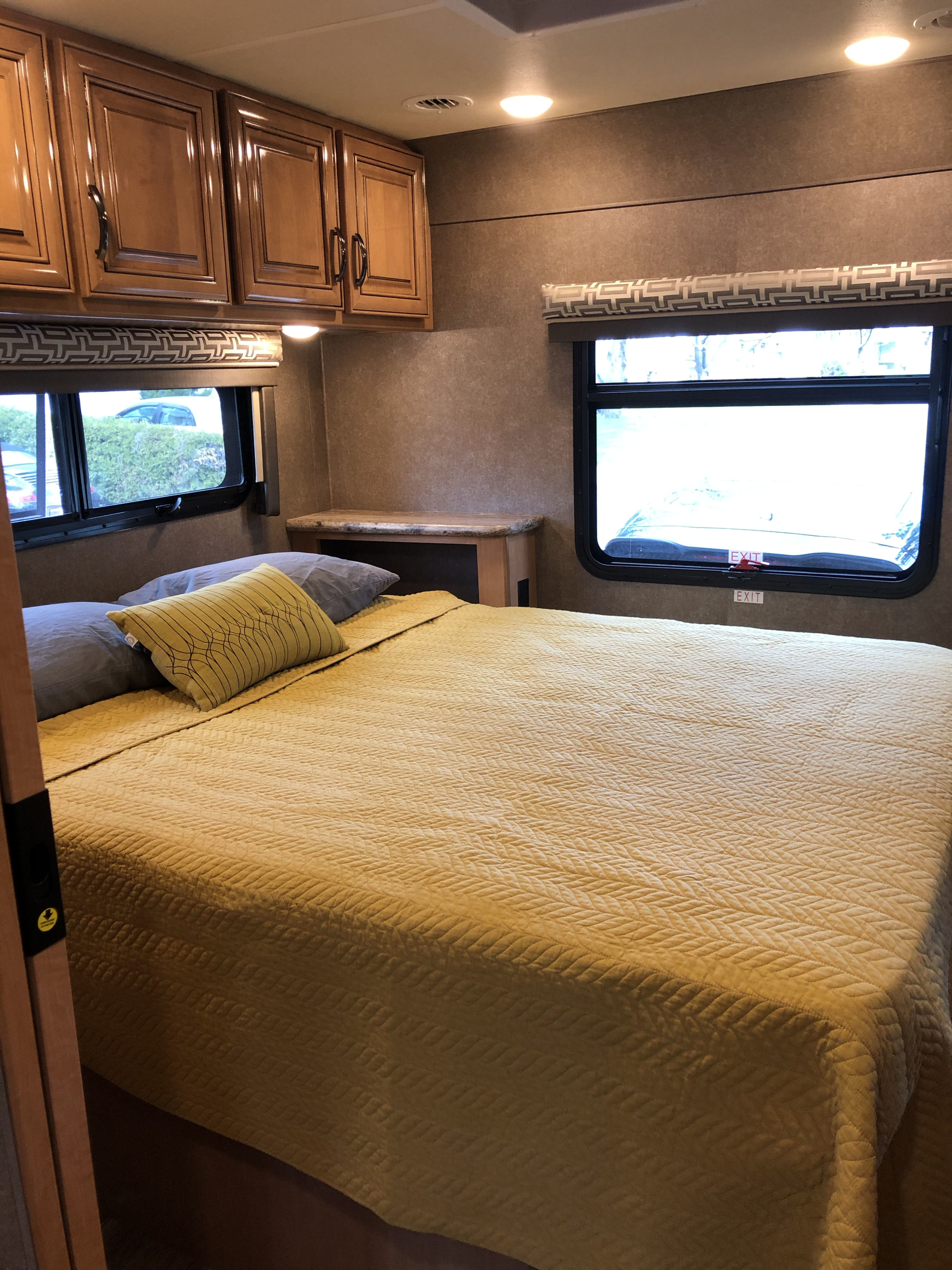 Cozy RV rental bedroom: yellow quilted bed, wooden cabinets, and windows with patterned shades.. Thor Quantum 2018