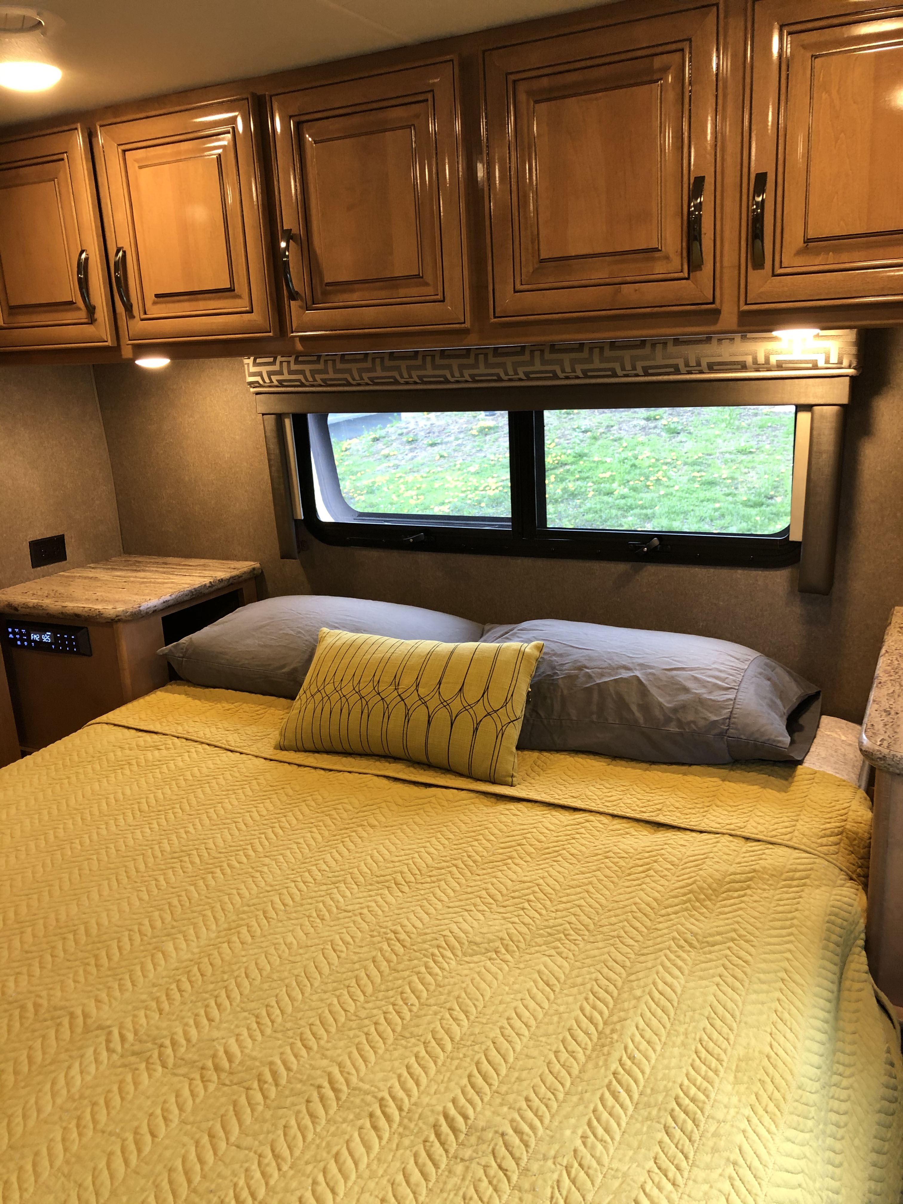 Inside an RV rental: a cozy bedroom with bright yellow quilted bedding, wood cabinets, and a scenic window view.. Thor Quantum 2018