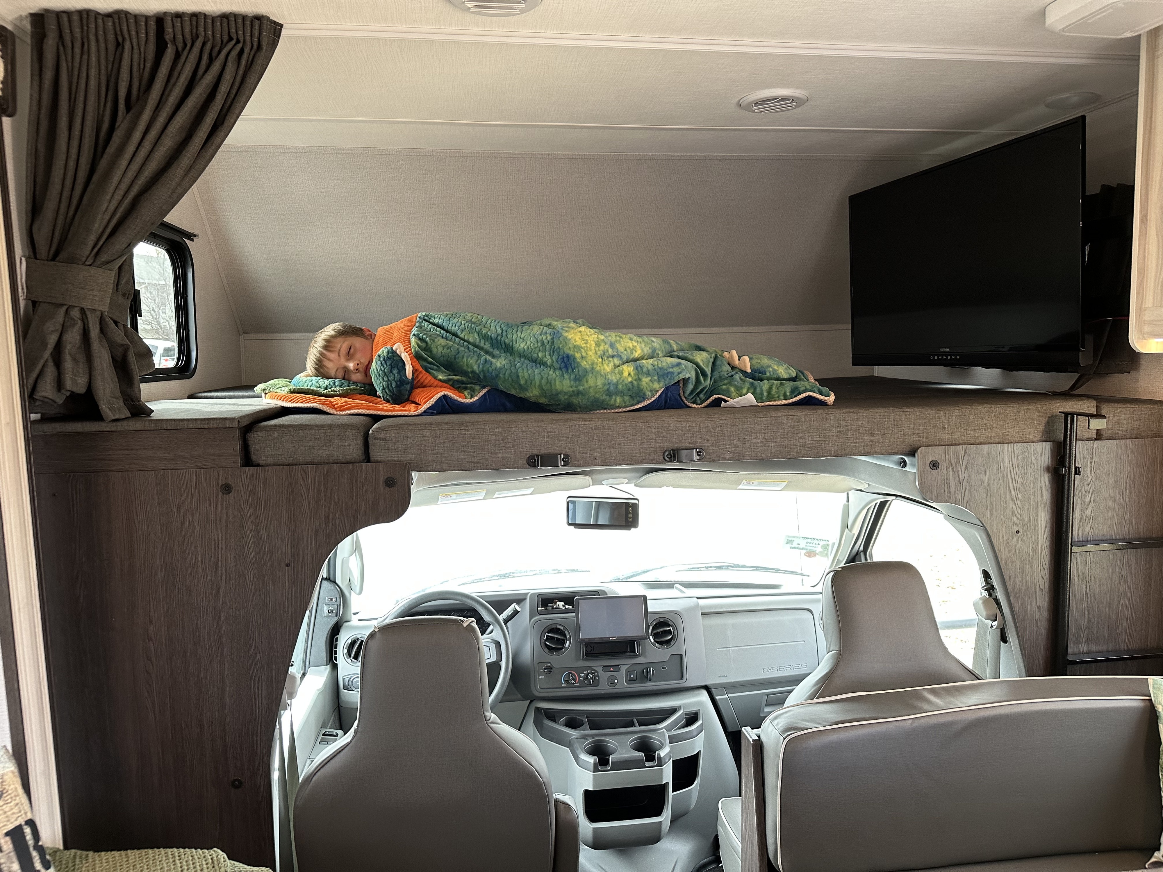 Child sleeps soundly in a cozy overhead bunk of this RV rental, with the driver's cab visible below.. Forest River Sunseeker 2023