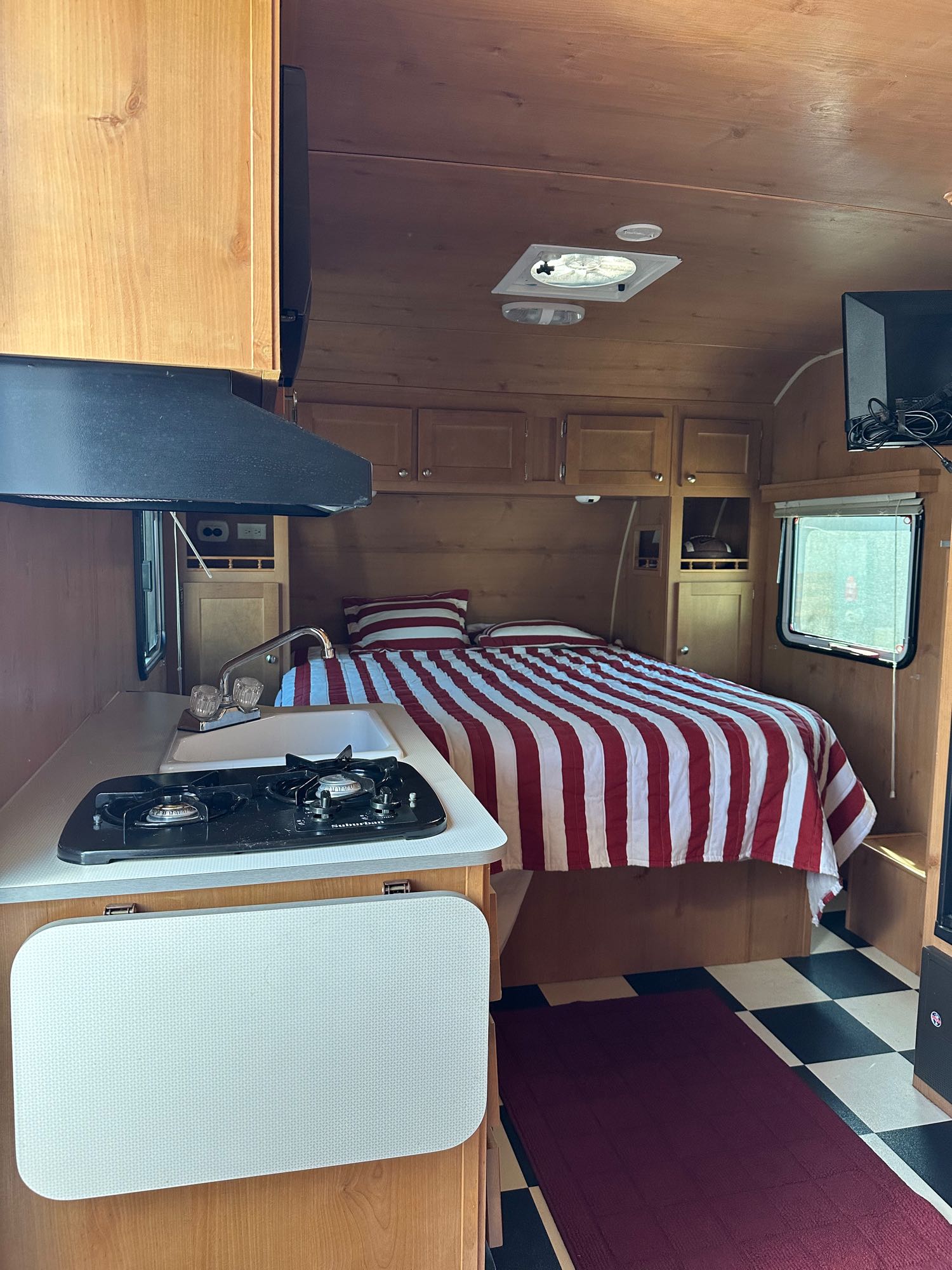 Inside a compact RV rental: full bed, kitchen with stove & sink, wood cabinets, TV, and checkered floor.. Riverside Travel Trailer Riverside Travel Trailer Trailer 2016