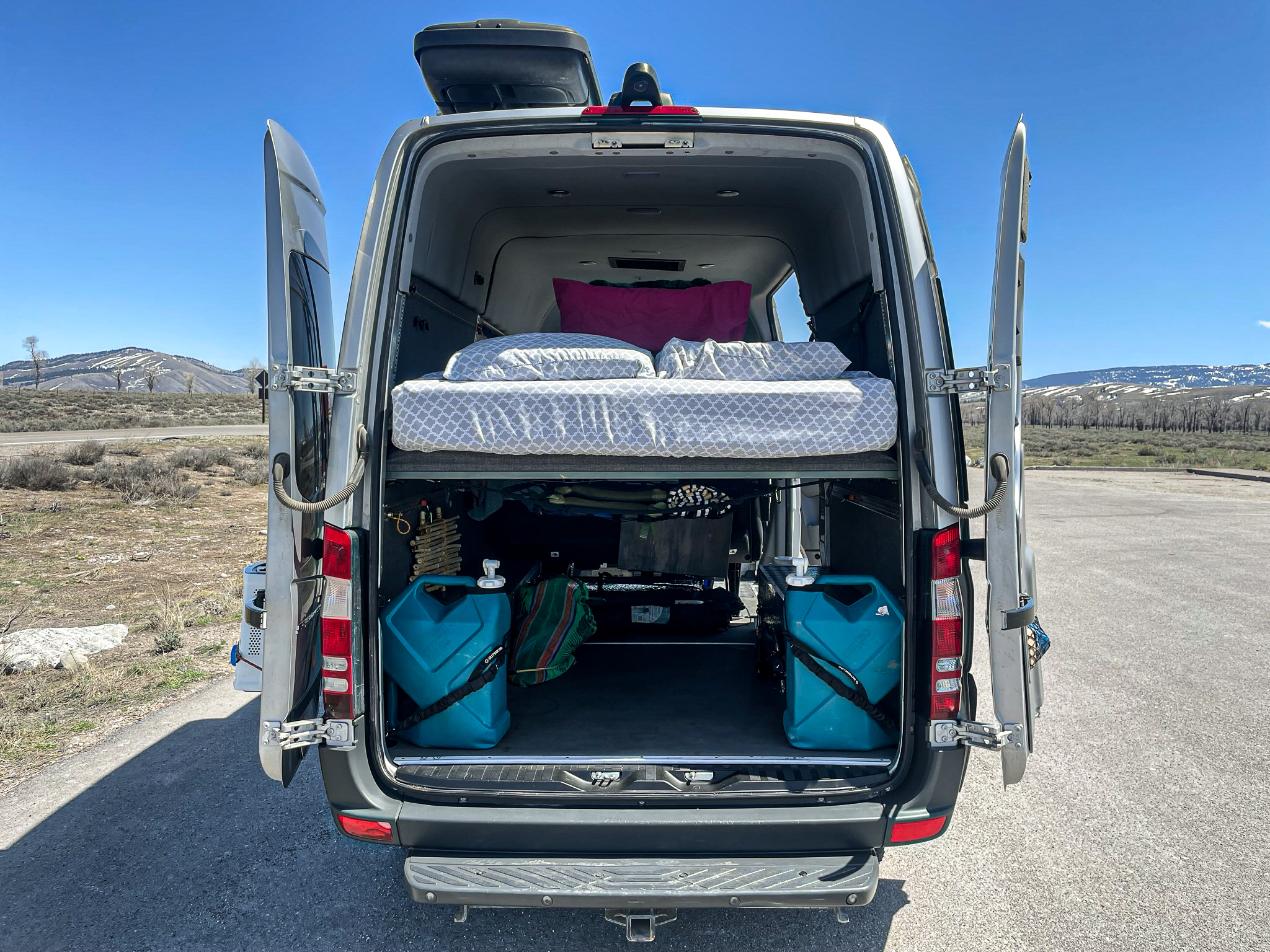 Open rear doors of an RV rental show a cozy bed, ample storage, and water containers, ready for adventure in the mountains.. Mercedes-Benz Sprinter 2016