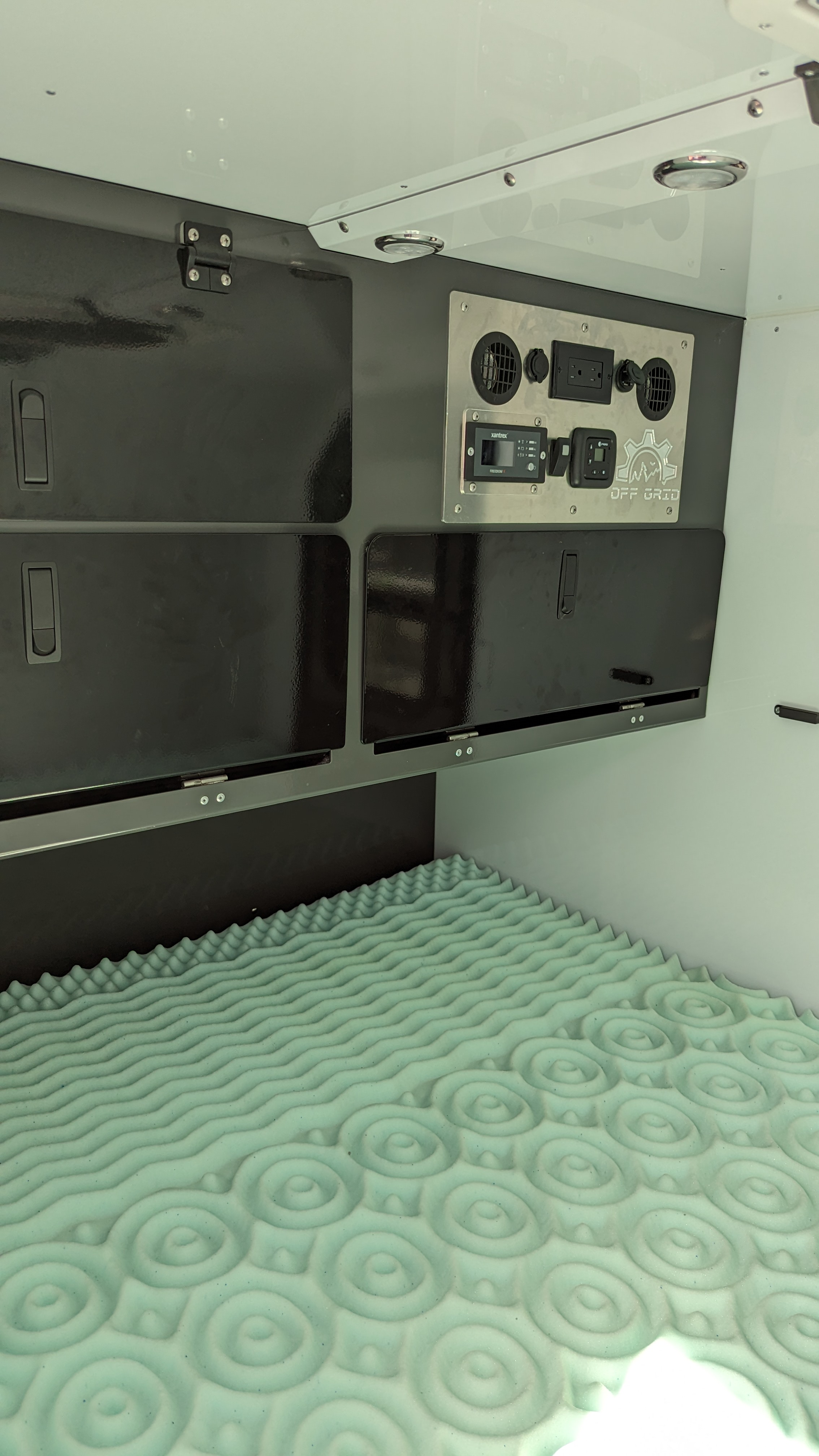 RV rental interior: black overhead storage, control panel with power outlets, and a textured foam mattress.. Off Grid Trailers Off Grid Trailers Trailer 2020