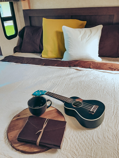 Cozy RV rental bedroom setup: a bed with pillows, a dark ukulele, journal, and mug on a wooden tray for relaxing.. Forest River Sunseeker 2019
