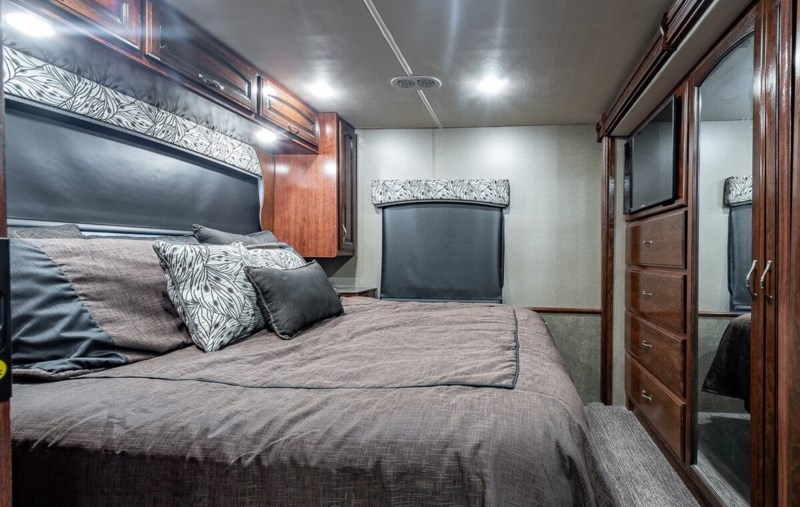 Cozy RV rental bedroom features a large bed with pillows, overhead cabinets, and wardrobe storage with a TV.. Holiday Rambler Vacationer XE 2018