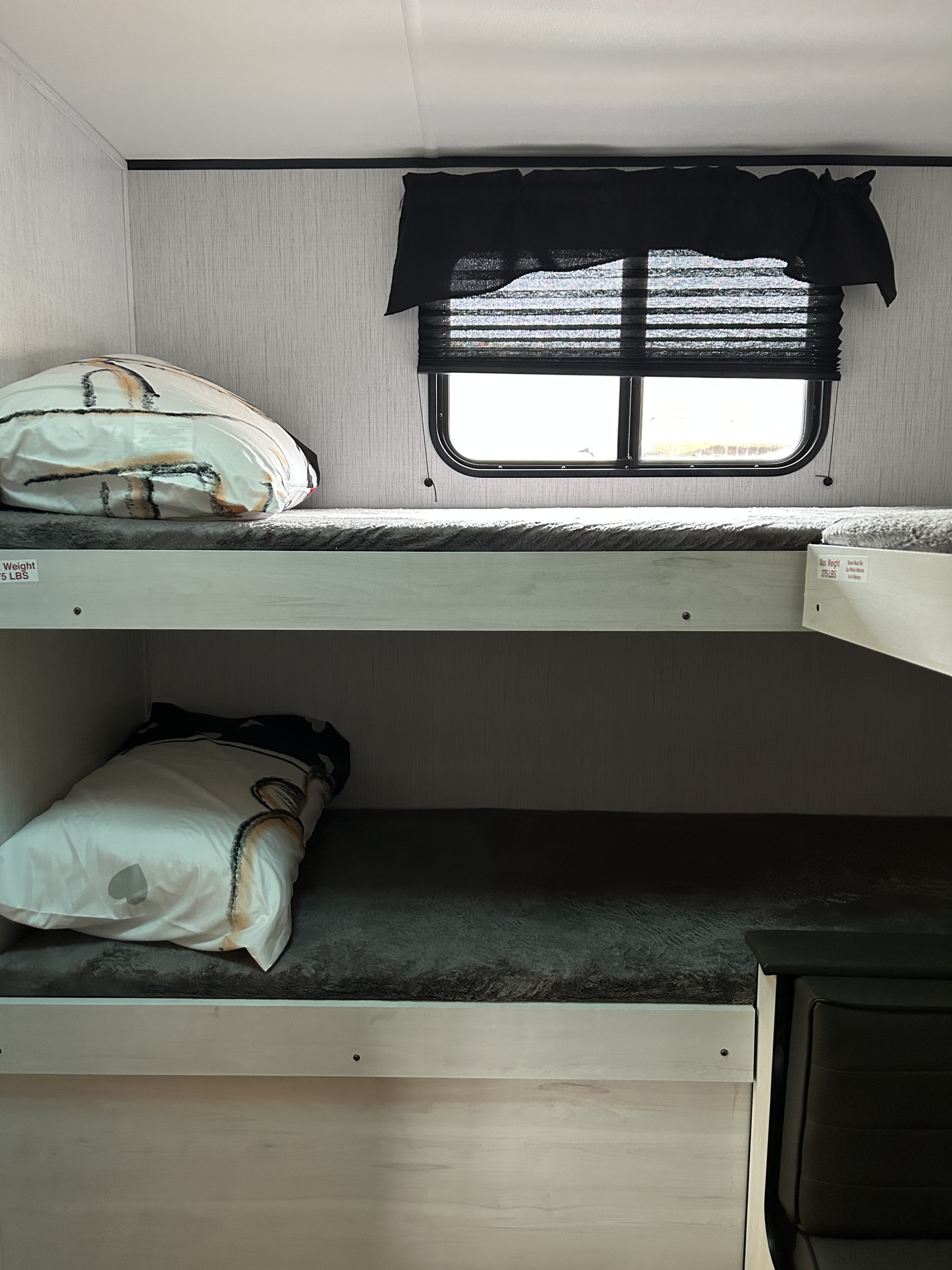 An rv rental's compact sleeping area features stacked bunk beds with soft pillows and a window with black privacy blinds.. Heartland RVs Trail Runner 2022